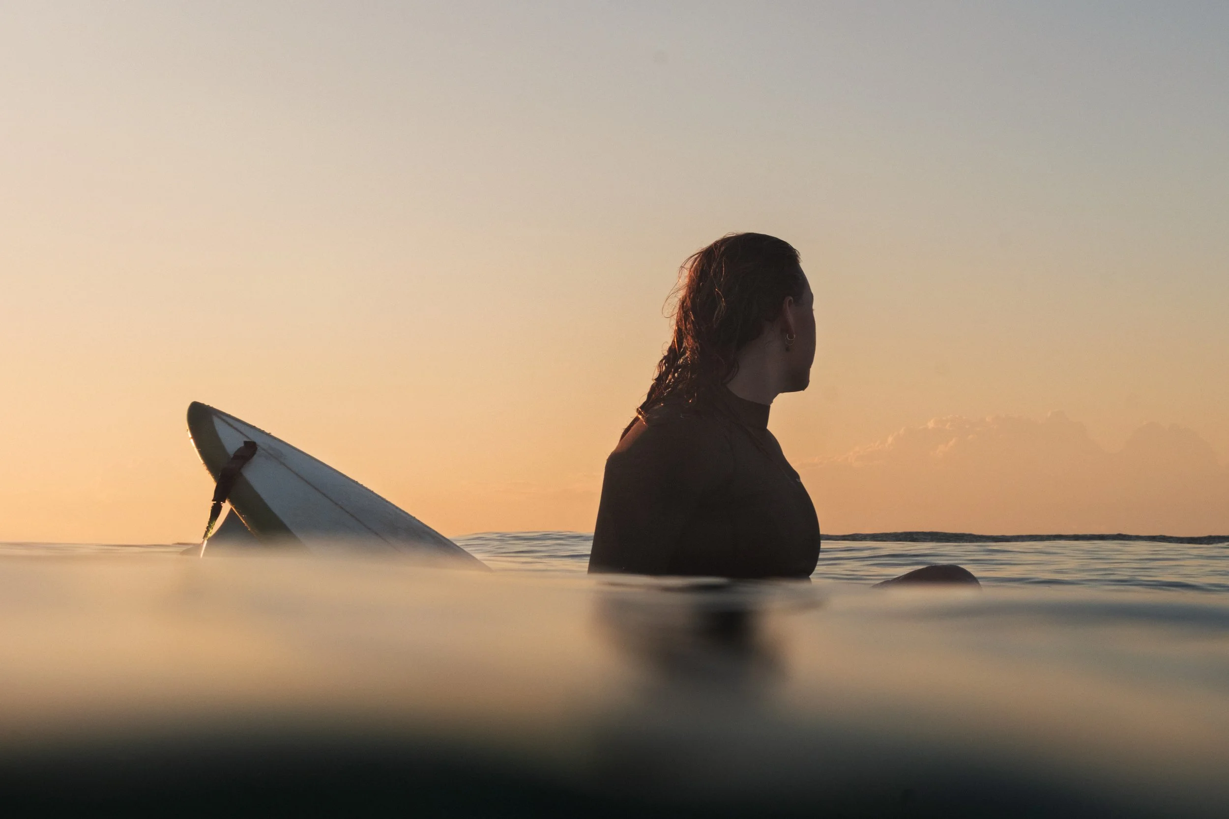 Person sitting in the water at sunset with surfboard partially submerged beside them.