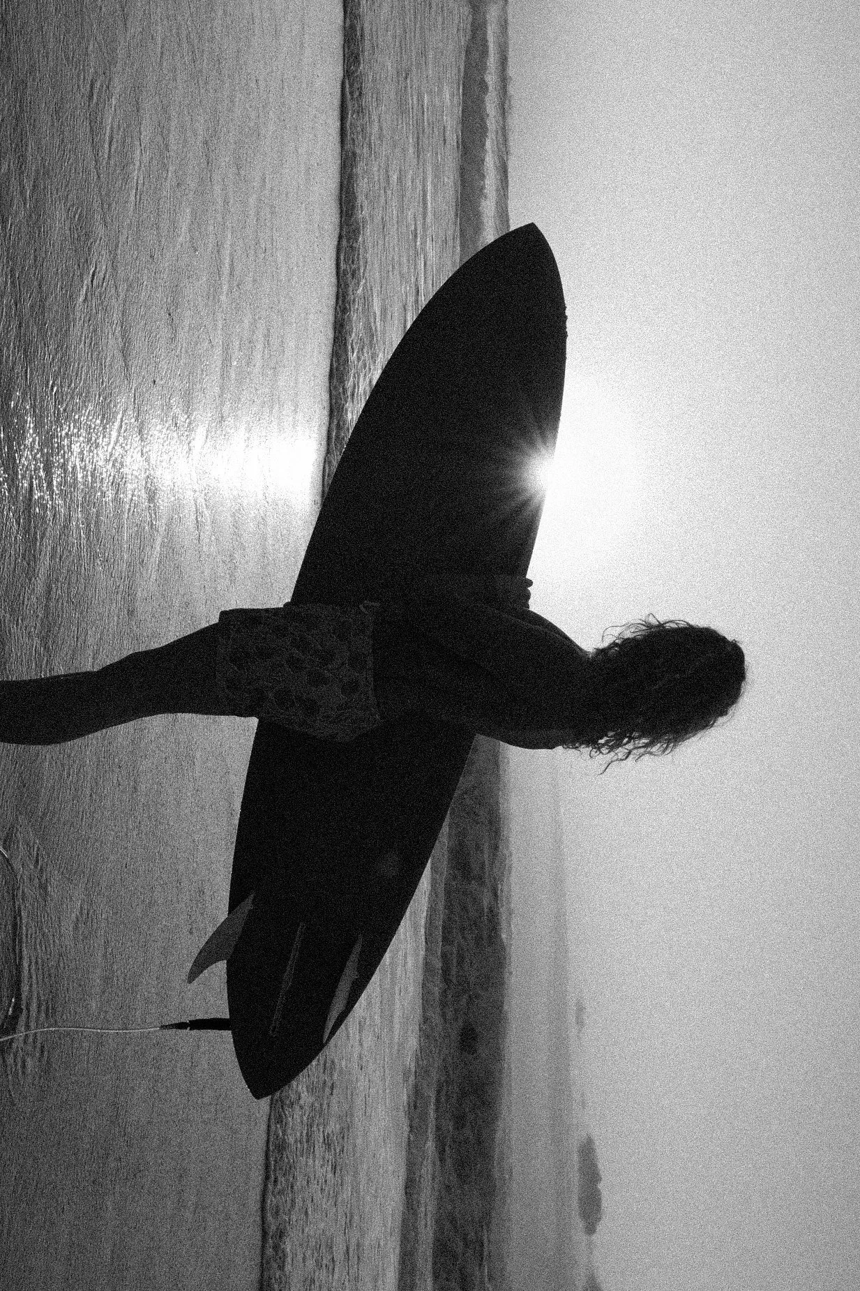 A person holding a surfboard on a beach at sunset, with waves in the background, in black and white.