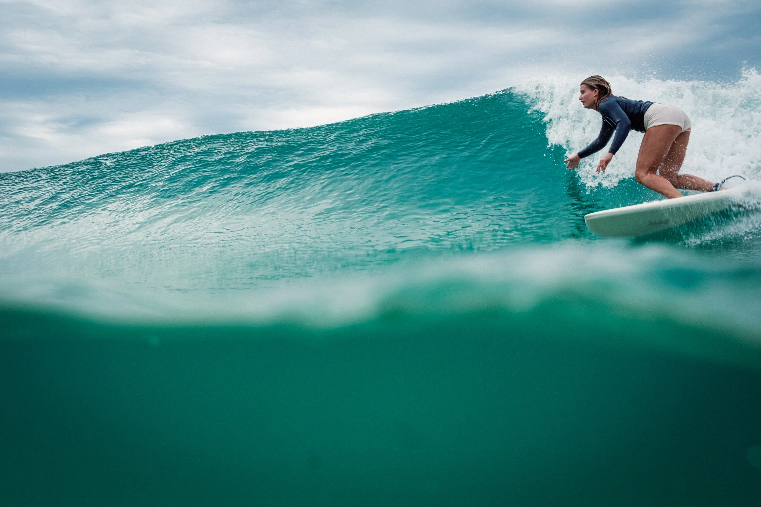 A woman surfing on a turquoise wave, wearing a black long-sleeved top and white shorts, with clouds in the sky above.