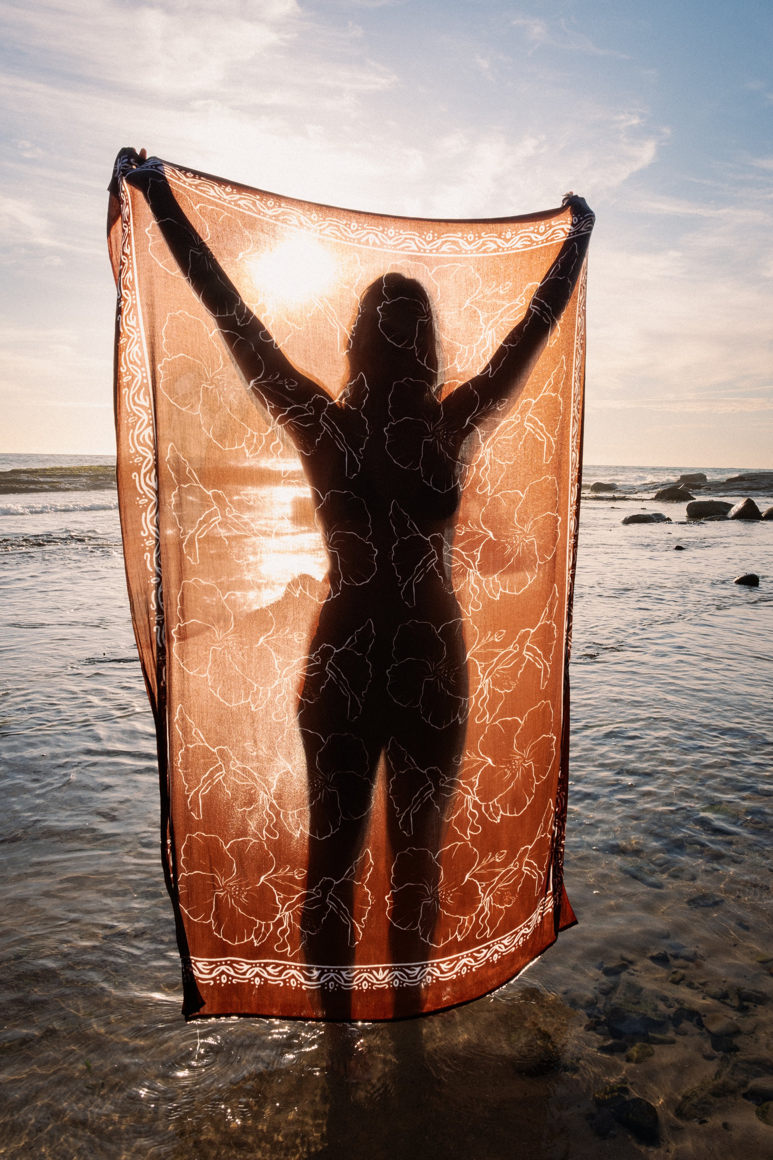 A silhouette of a woman holding a patterned cloth at the beach during sunset.