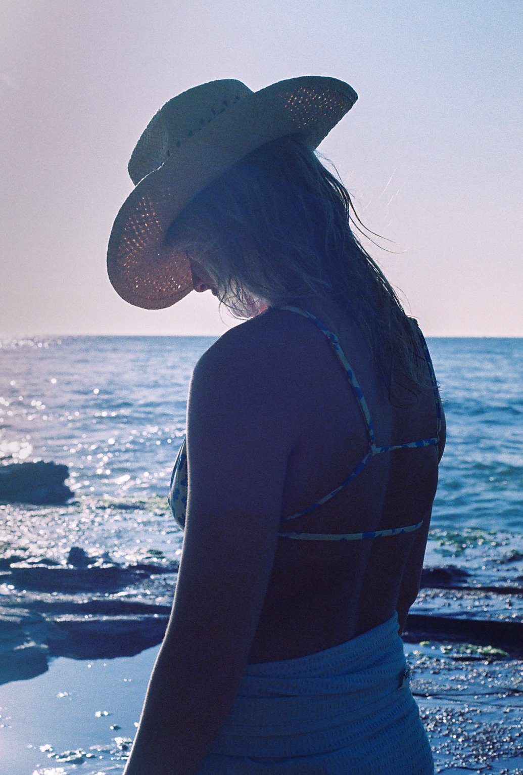 A woman standing on a beach wearing a straw hat, a swimsuit with straps, and a towel wrapped around her waist, facing the ocean during sunset.
