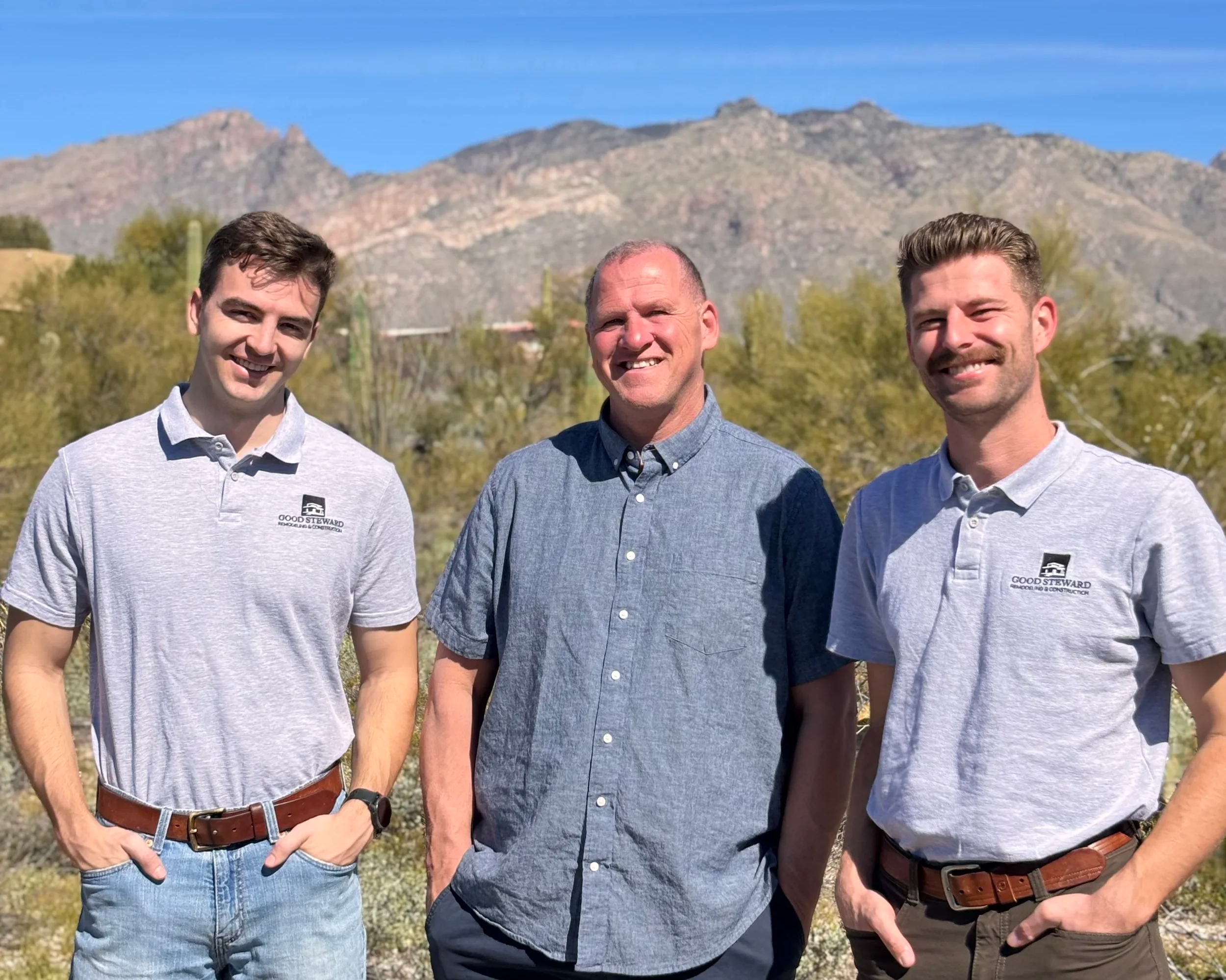 Three men standing outdoors with mountains in the background, smiling at the camera.