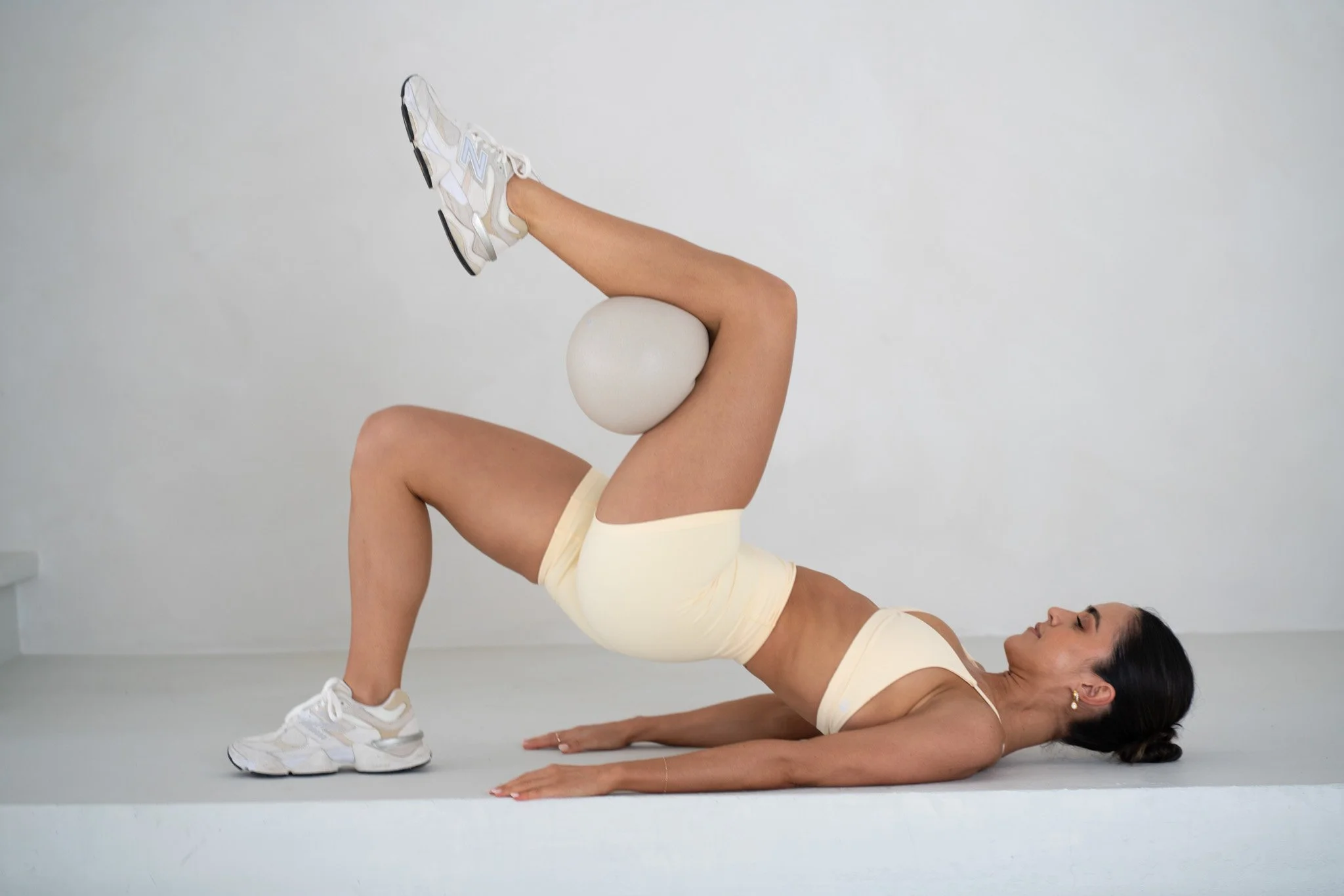Woman lying on her back doing a pilates exercise with a pilates ball placed between her knees and one foot raised with a sneaker, against a plain white background.