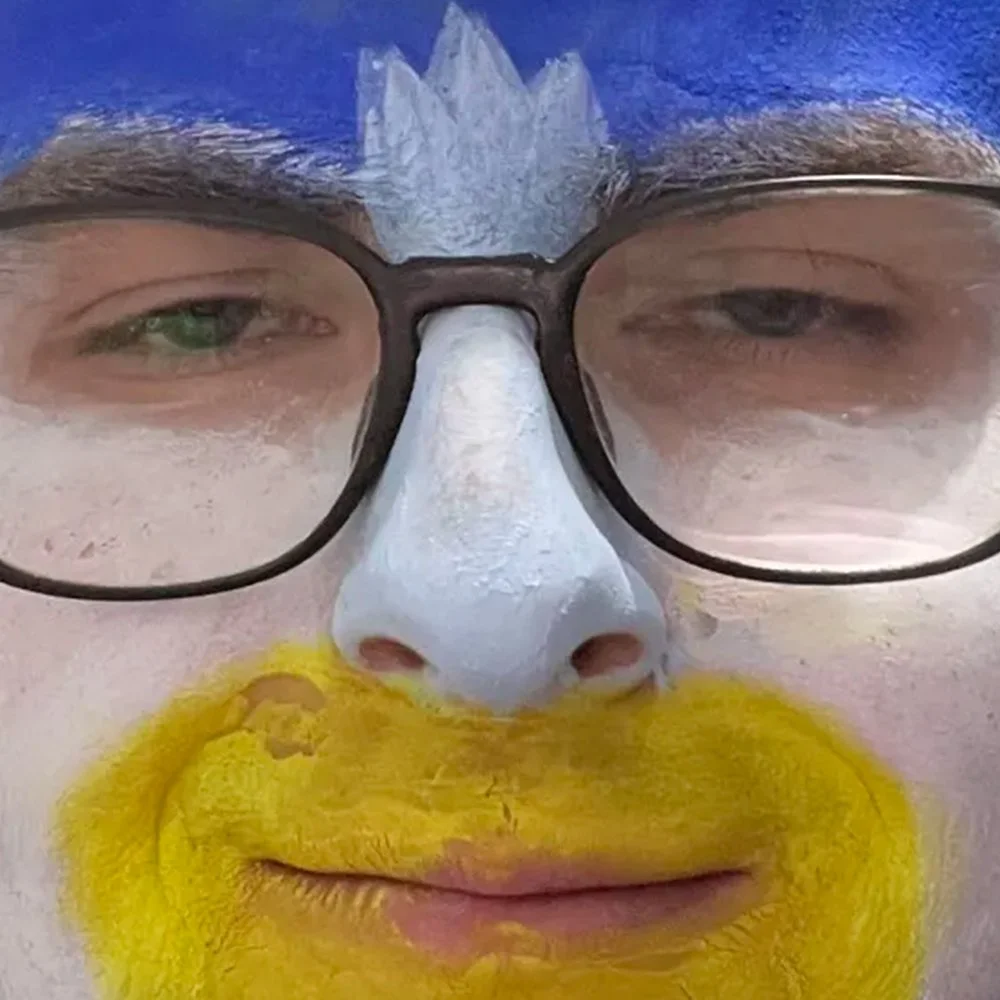 A close-up of a person's face with a painted yellow beard, white nose, and blue crown, wearing black-rimmed glasses.
