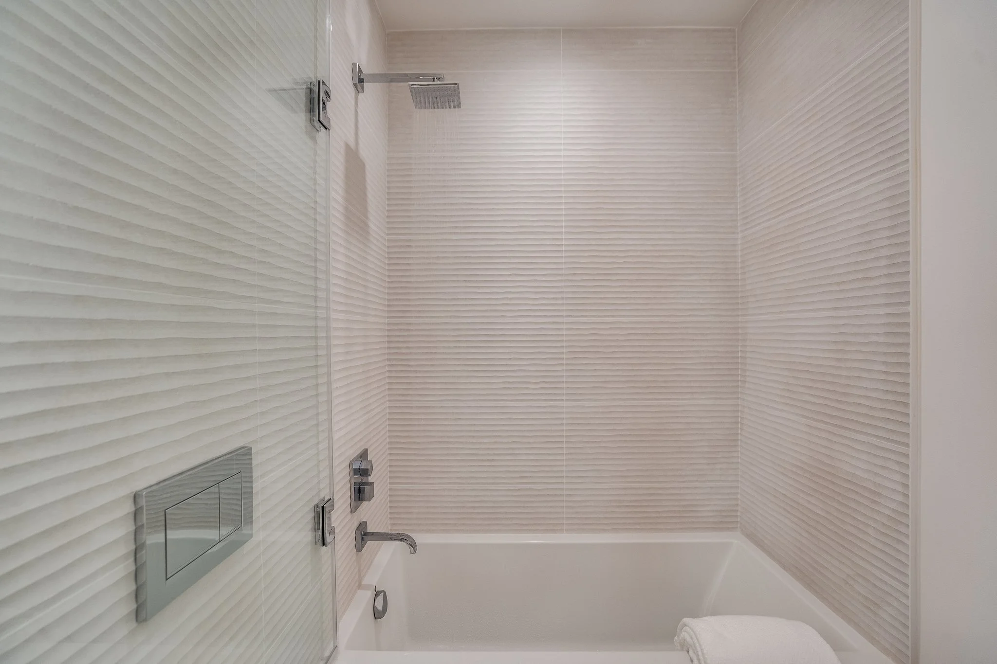 Modern bathroom with textured beige wall tiles, a glass shower enclosure, a rainfall shower head, a built-in toilet flush button, and a white bathtub with a towel on its edge.