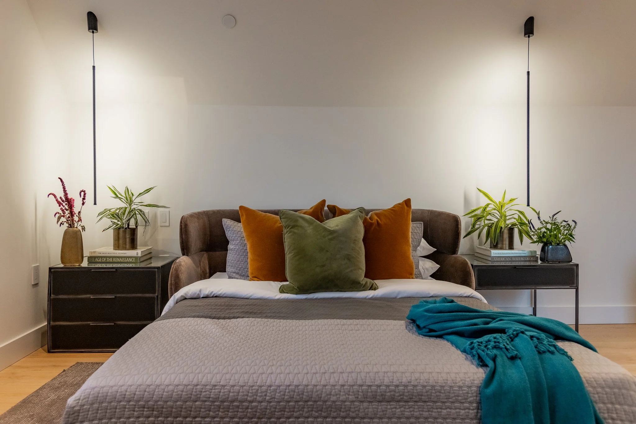 Modern bedroom with a bed, orange and green accent pillows, gray bedspread, blue throw blanket, two black nightstands with potted plants and books, white walls, two wall-mounted lamps, hardwood floor.