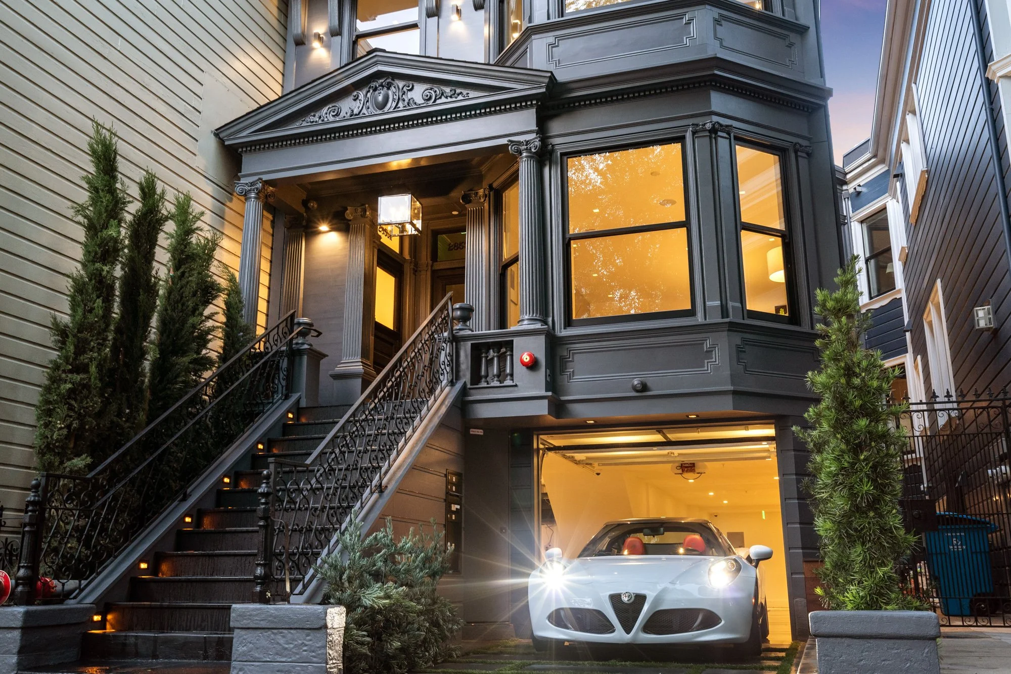 A black multi-story house with ornate architectural details, a staircase with metal railings on the left, and a silver Alfa Romeo sports car parked in the garage. The house has large windows with warm interior lighting, and there are trees and plants