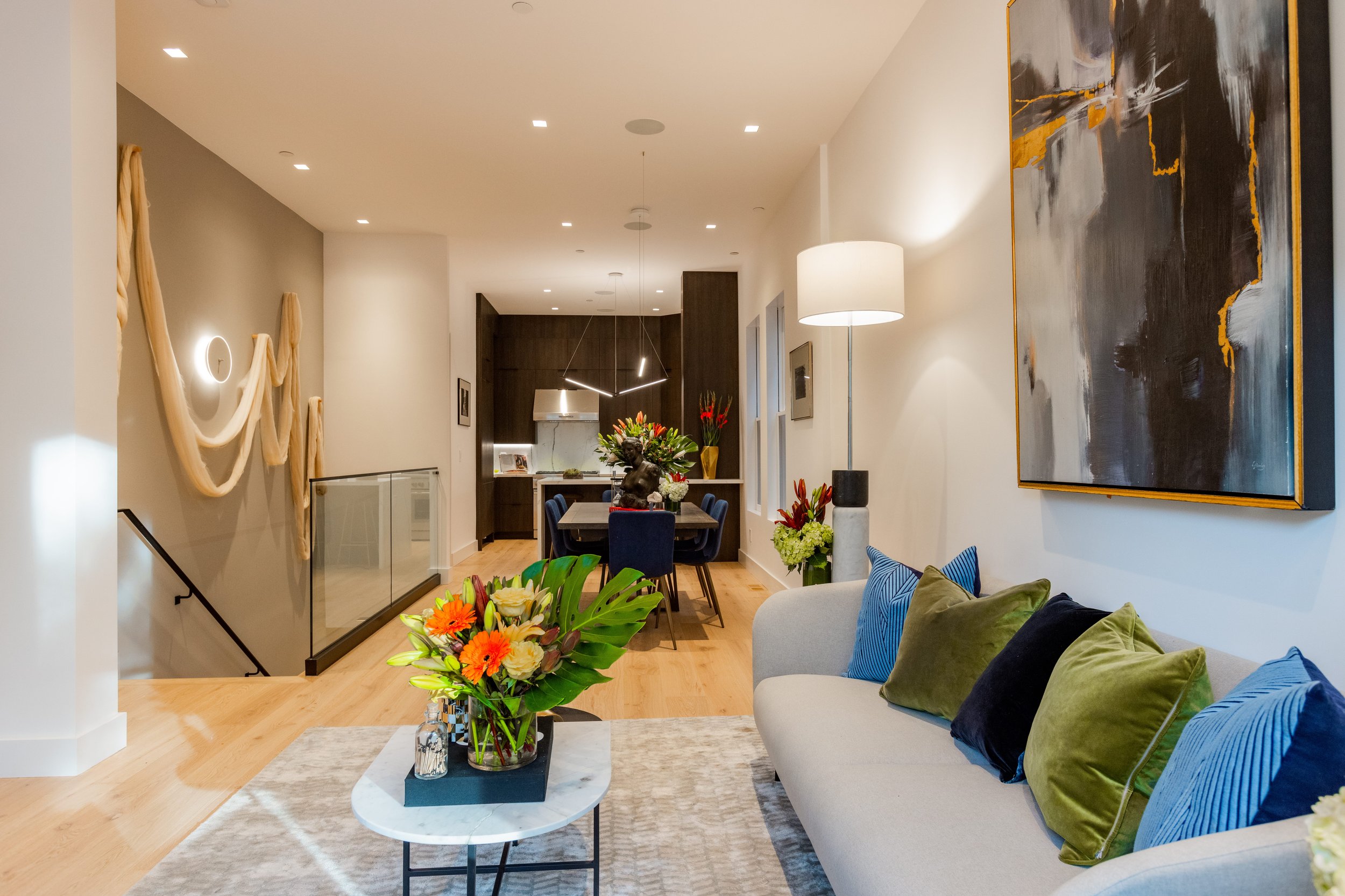 Modern living room with a white sofa, colorful pillows, a coffee table with a floral arrangement, abstract art on the wall, and a dining area and kitchen in the background.