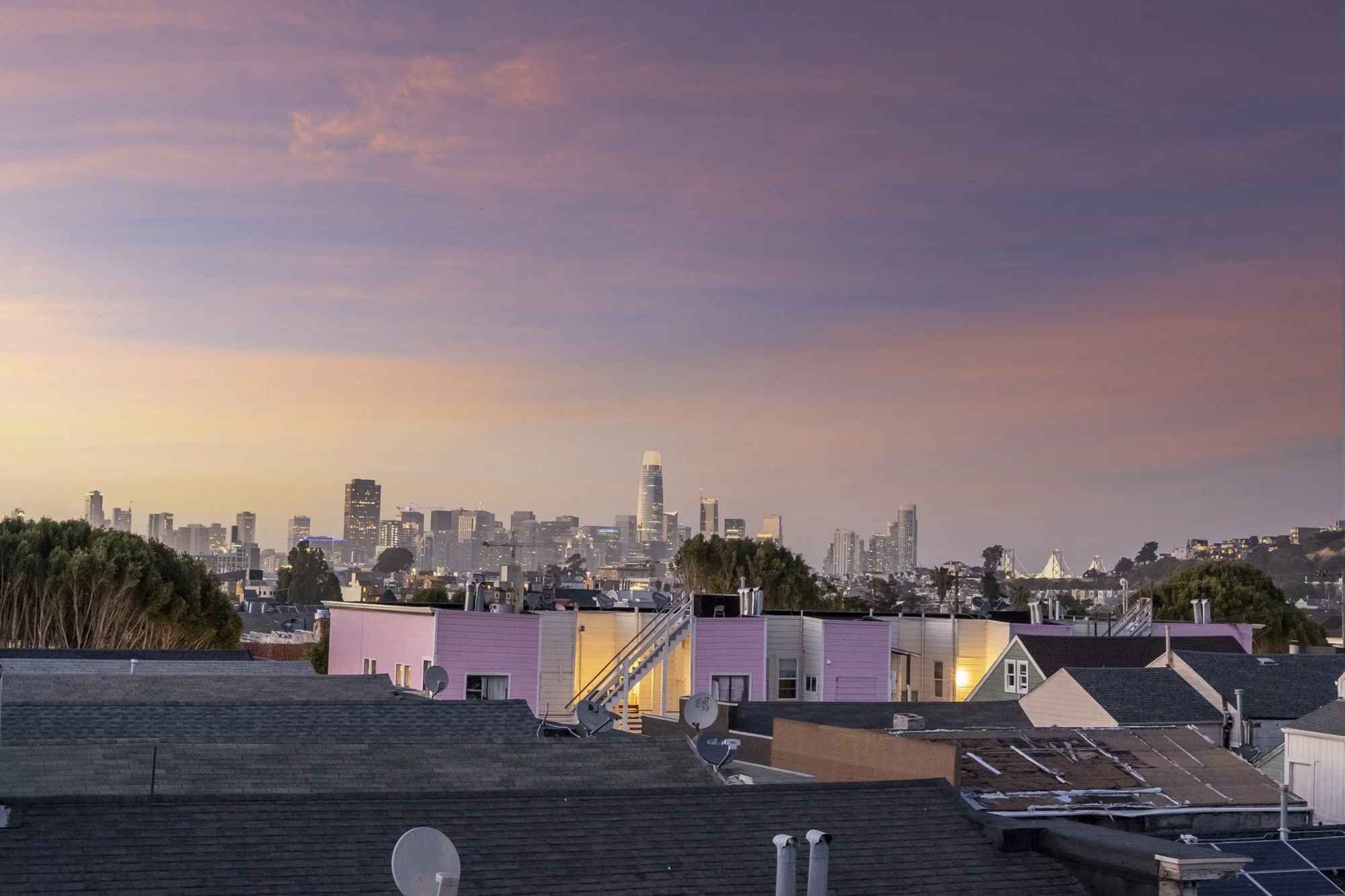 City skyline at sunset with tall buildings in the background and residential houses with satellite dishes in the foreground.