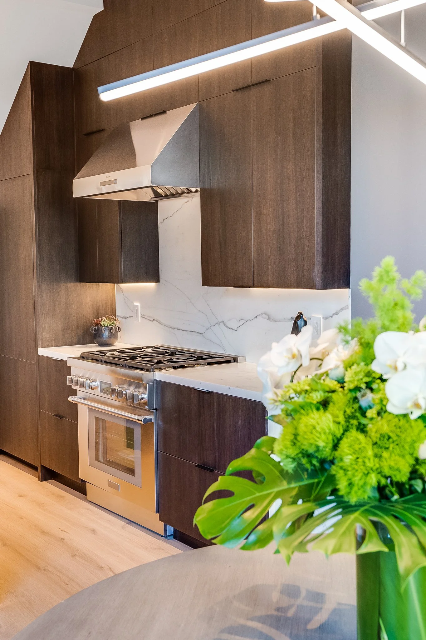 Modern kitchen with dark wood cabinets, a stainless steel range hood, a stainless steel oven, and a white marble backsplash. A bouquet of white flowers and green plants on a table in the foreground.