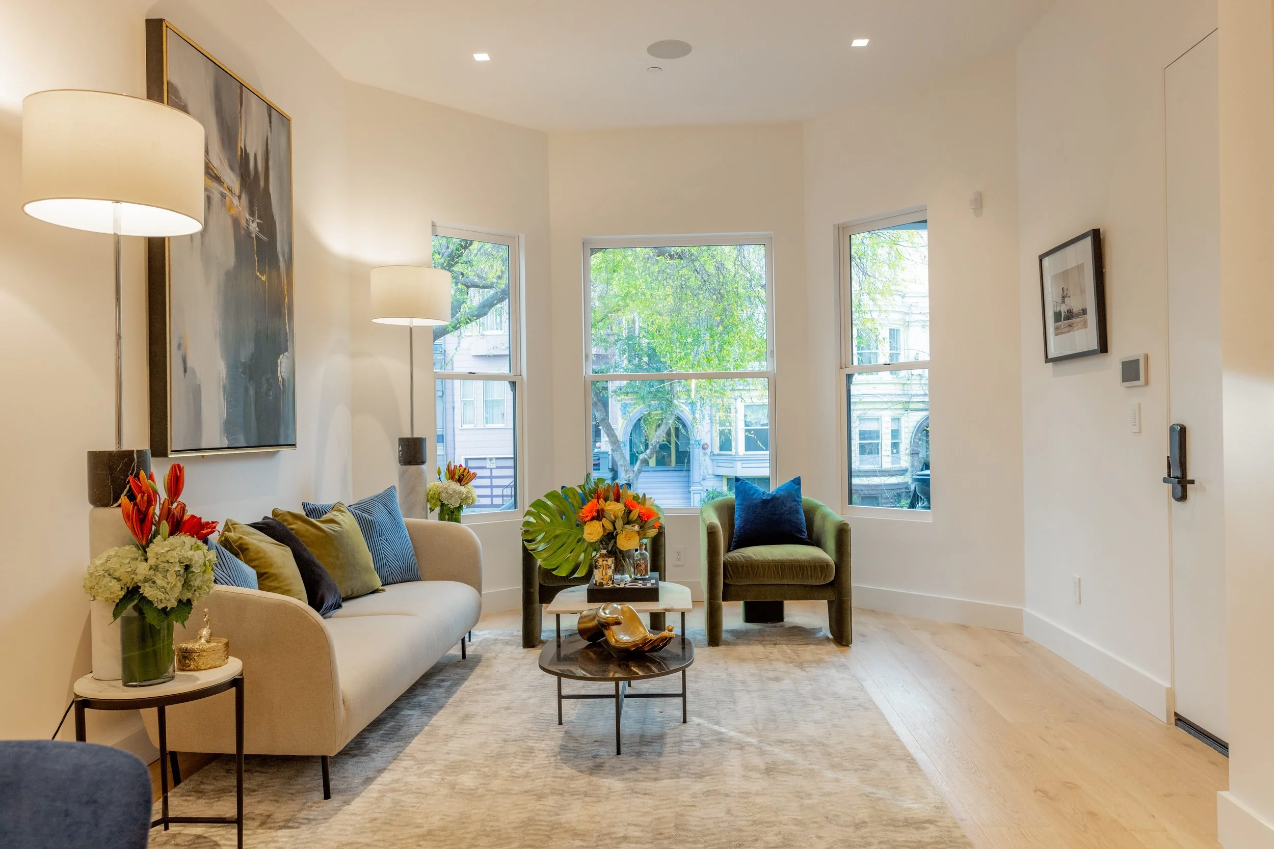 A modern living room with a beige sofa, green accent chair, and floor lamps, decorated with colorful flowers and wall art, illuminated by natural light from large windows.