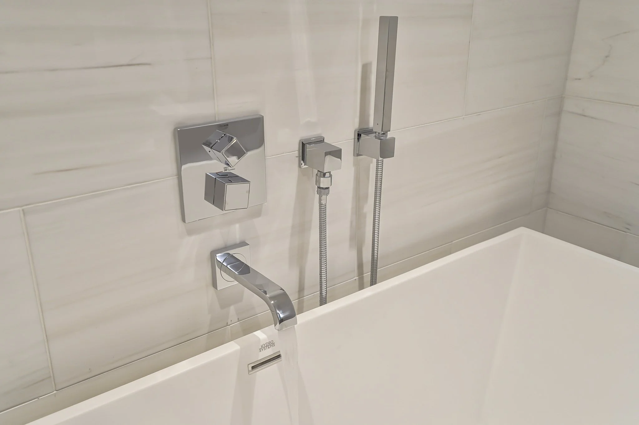 Modern bathtub with chrome fixtures including a faucet, two control knobs, and a handheld shower head with hose, installed on a tiled wall.