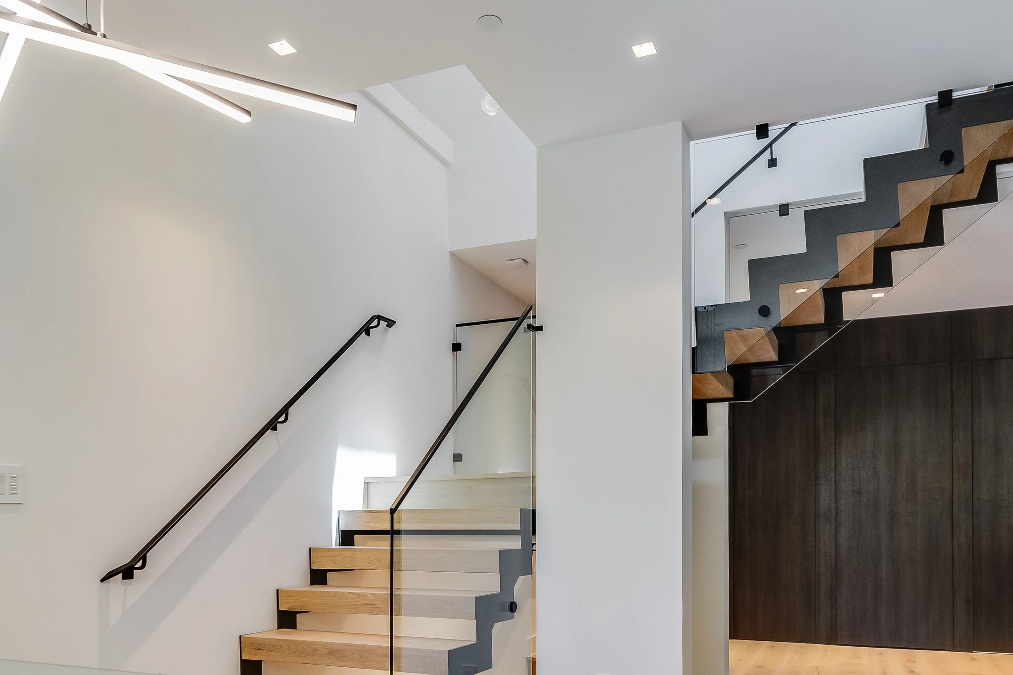 Interior view of a modern staircase with light wood steps, black handrails, and a dark wall, illuminated by ceiling lights in a minimalist home.