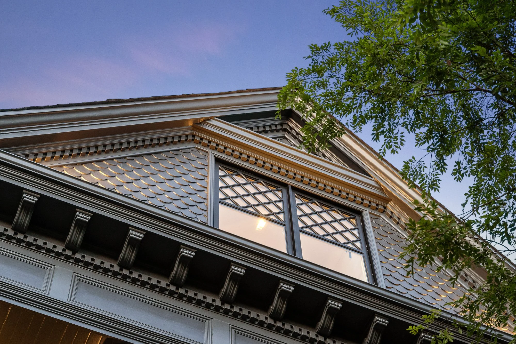 Close-up of a gabled roof with decorative shingles, ornate trim, and a large window reflecting the sky, with tree branches and green leaves in the foreground.