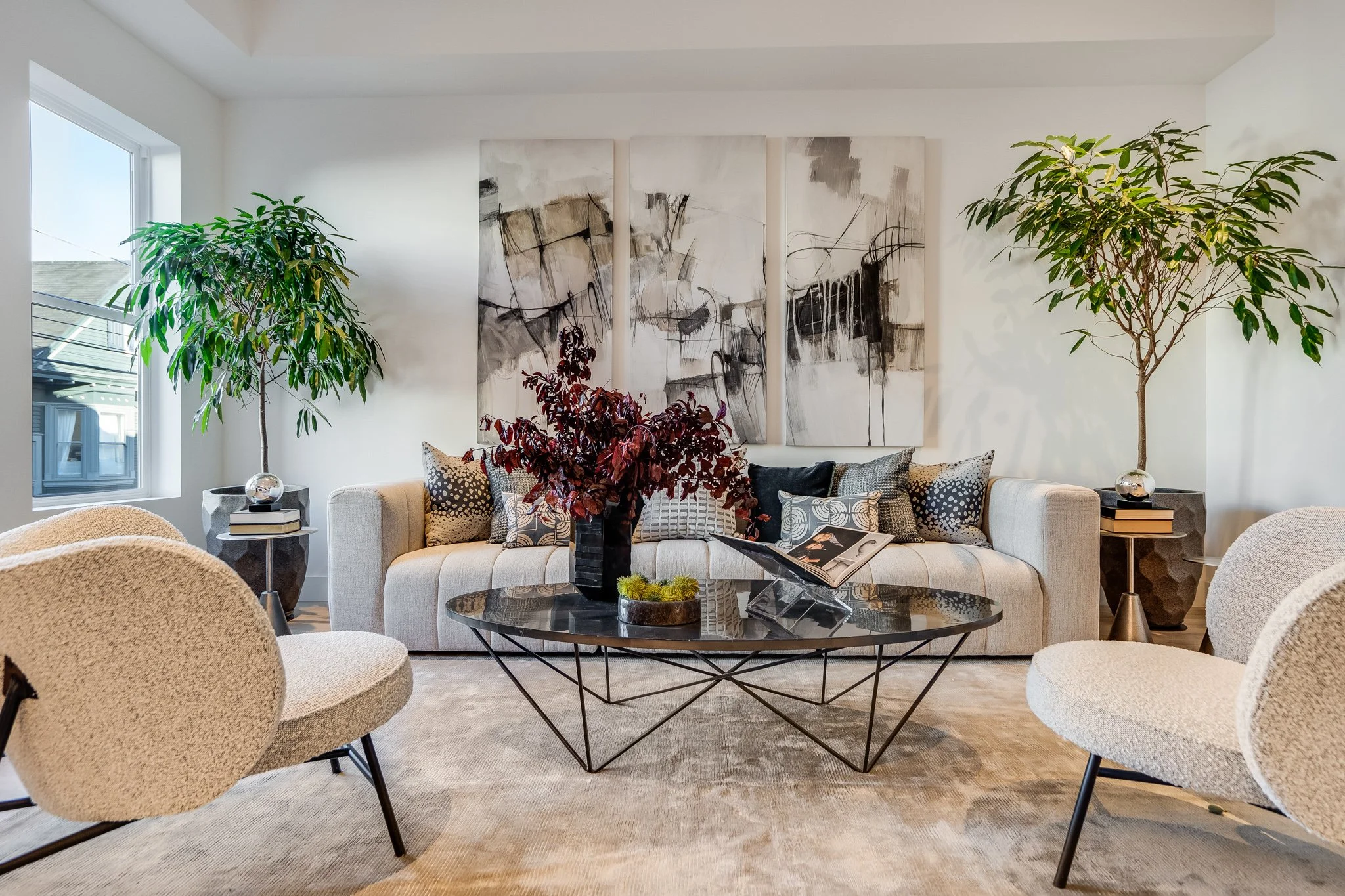 Modern living room with a beige sofa, black glass coffee table, white textured chairs, large plants in the corners, and abstract artwork behind the sofa.