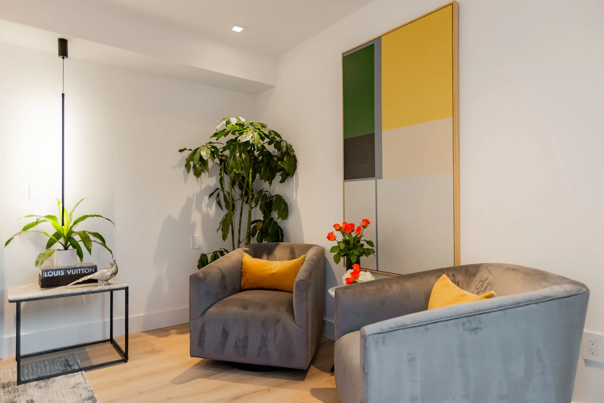 Living room corner with two grey velvet armchairs with yellow pillows, a small white side table with potted plants, a tall green leafy plant, a colorful abstract painting on the wall, and a light-colored wooden floor.