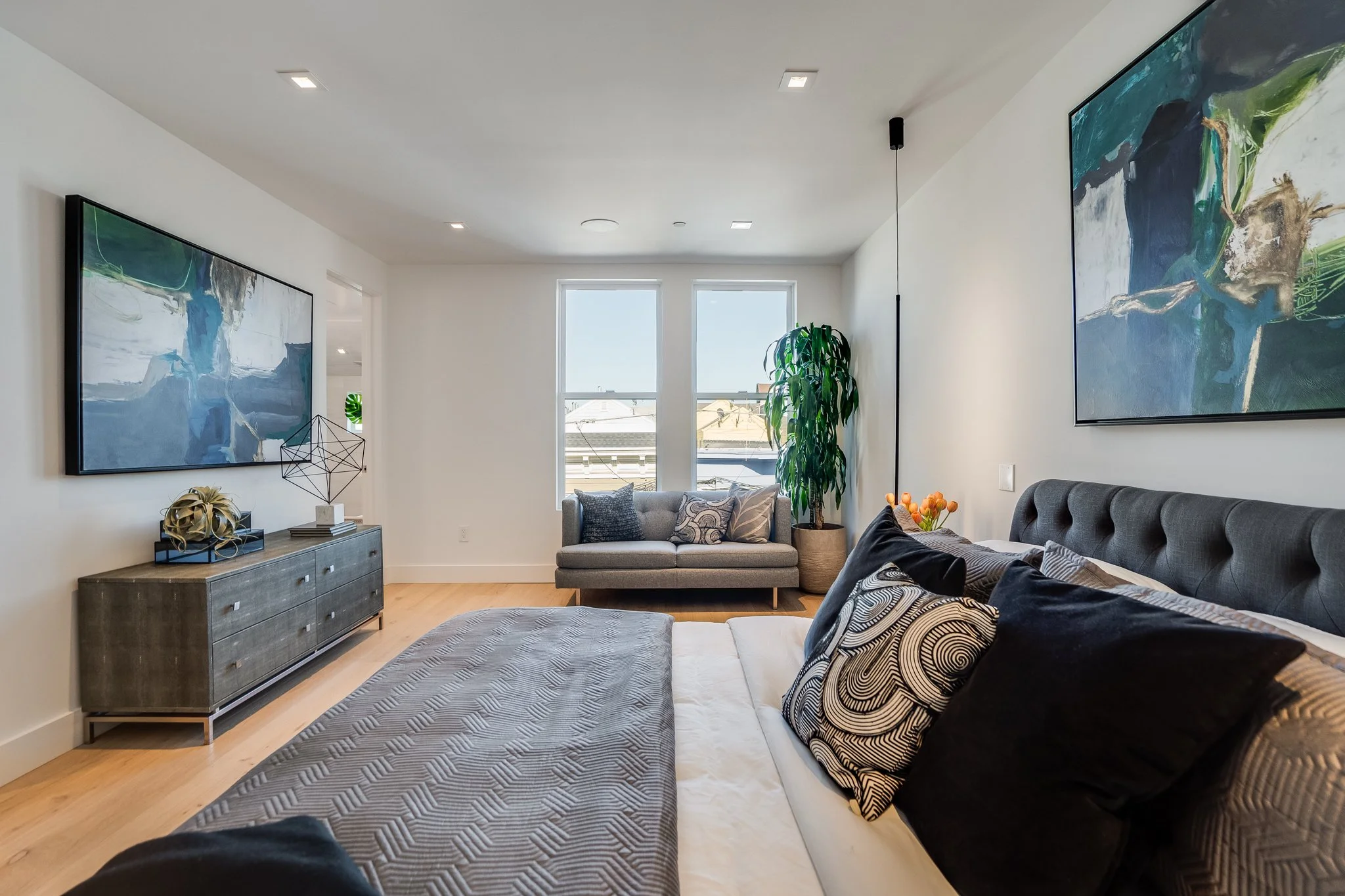 Modern living room with a bed, gray headboard, black and patterned pillows, a gray dresser with decorative items, a gray sofa under two windows, a large artwork on the wall, a potted plant, and a small side table with orange flowers.