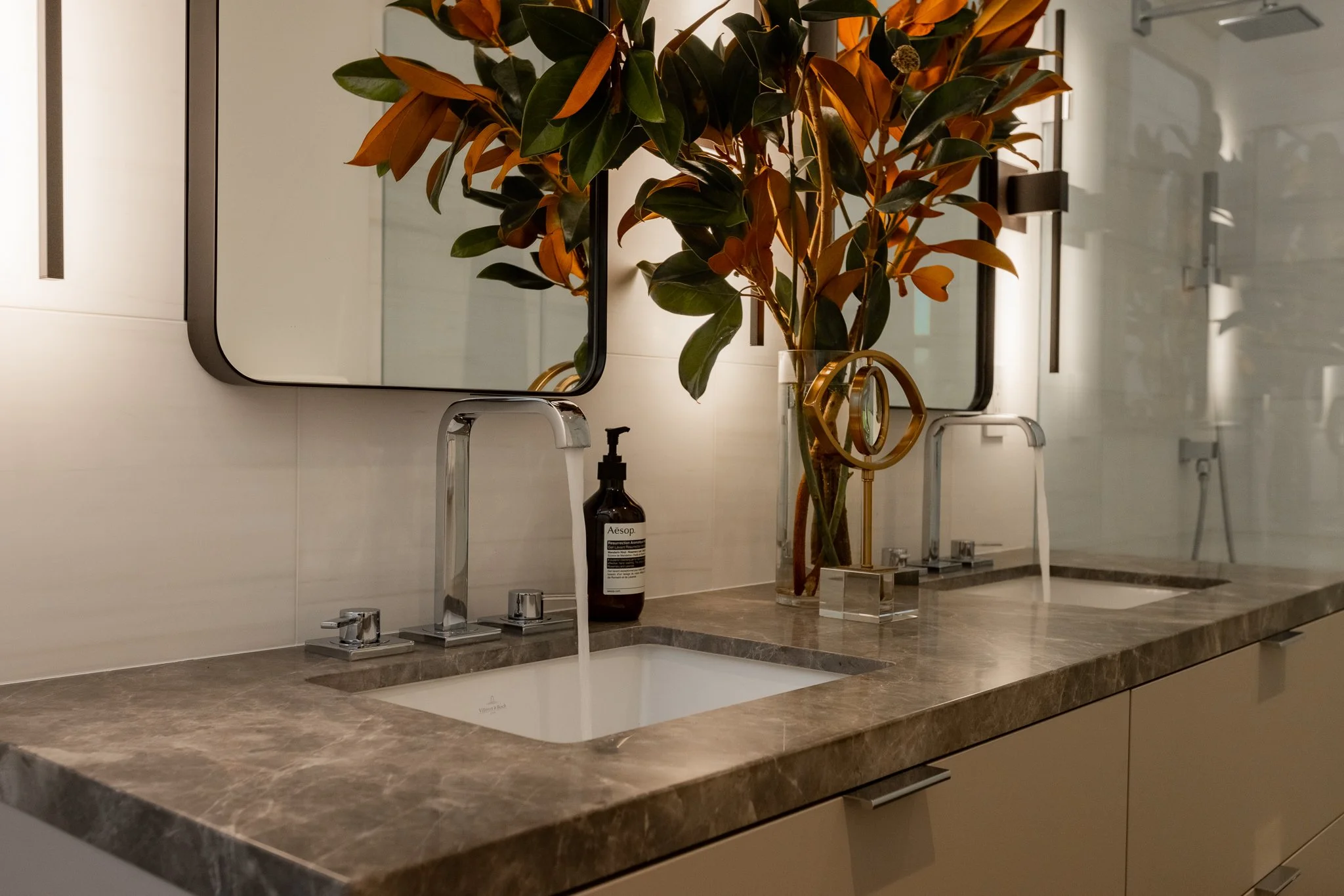 Bathroom vanity with two sinks, marble countertop, two mirrors, a potted plant with large green leaves, a black soap bottle, and a gold decorative object.