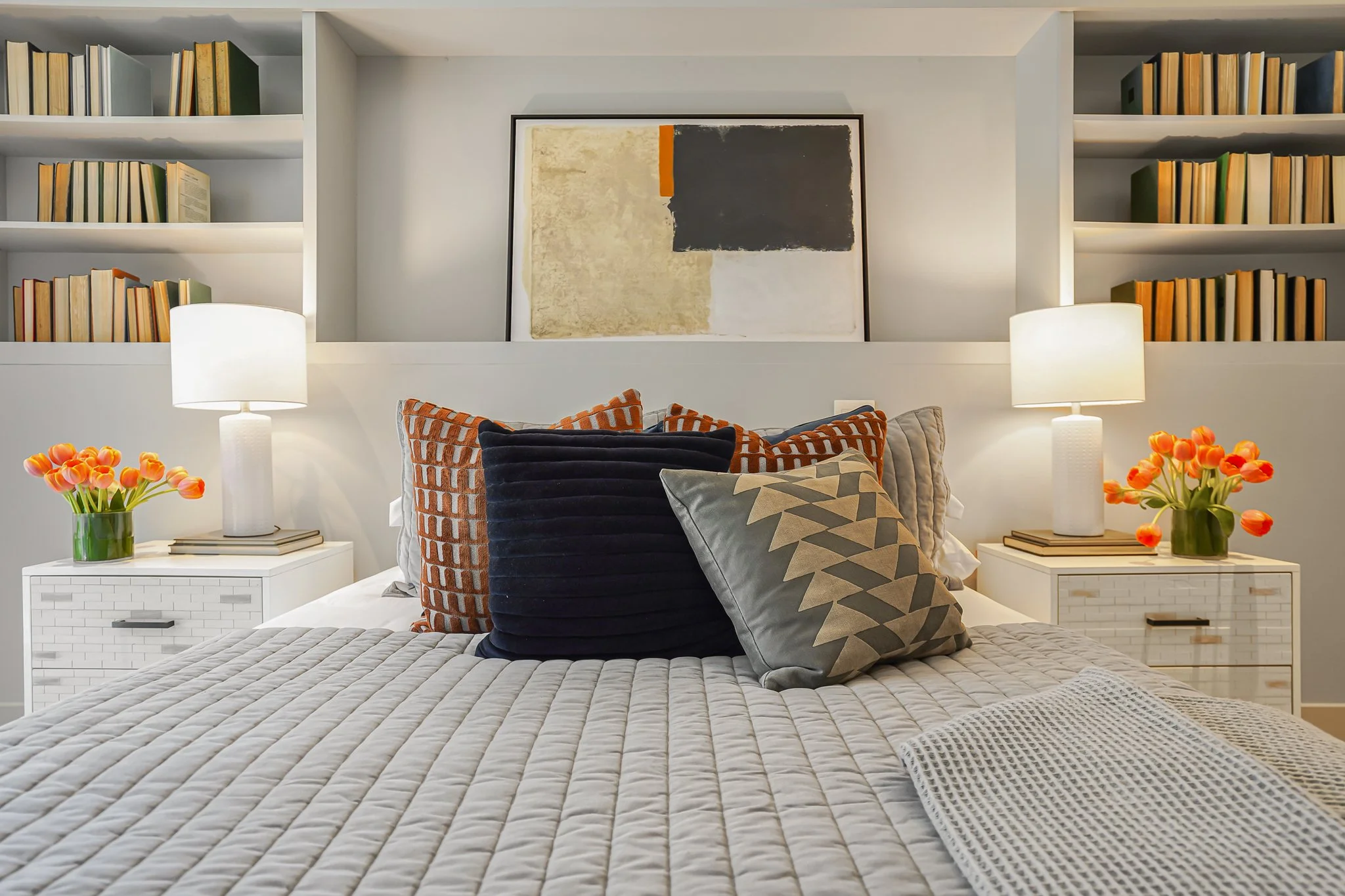 A neatly made bed with beige quilted bedding and decorative pillows in front of a white headboard, flanked by white nightstands with lamps and orange tulip vases. Behind the bed are built-in bookshelves with books and a framed abstract art piece abov