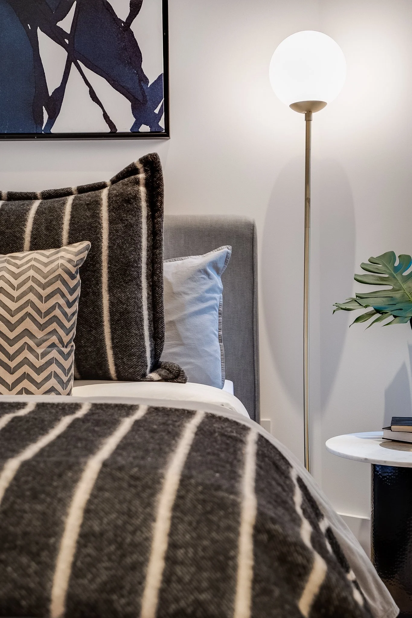 A bedroom with a bed featuring a gray upholstered headboard, striped and chevron-patterned pillows, a tall floor lamp with a round white shade, a white nightstand with a stack of books, and a plant with large green leaves, against a white wall with b