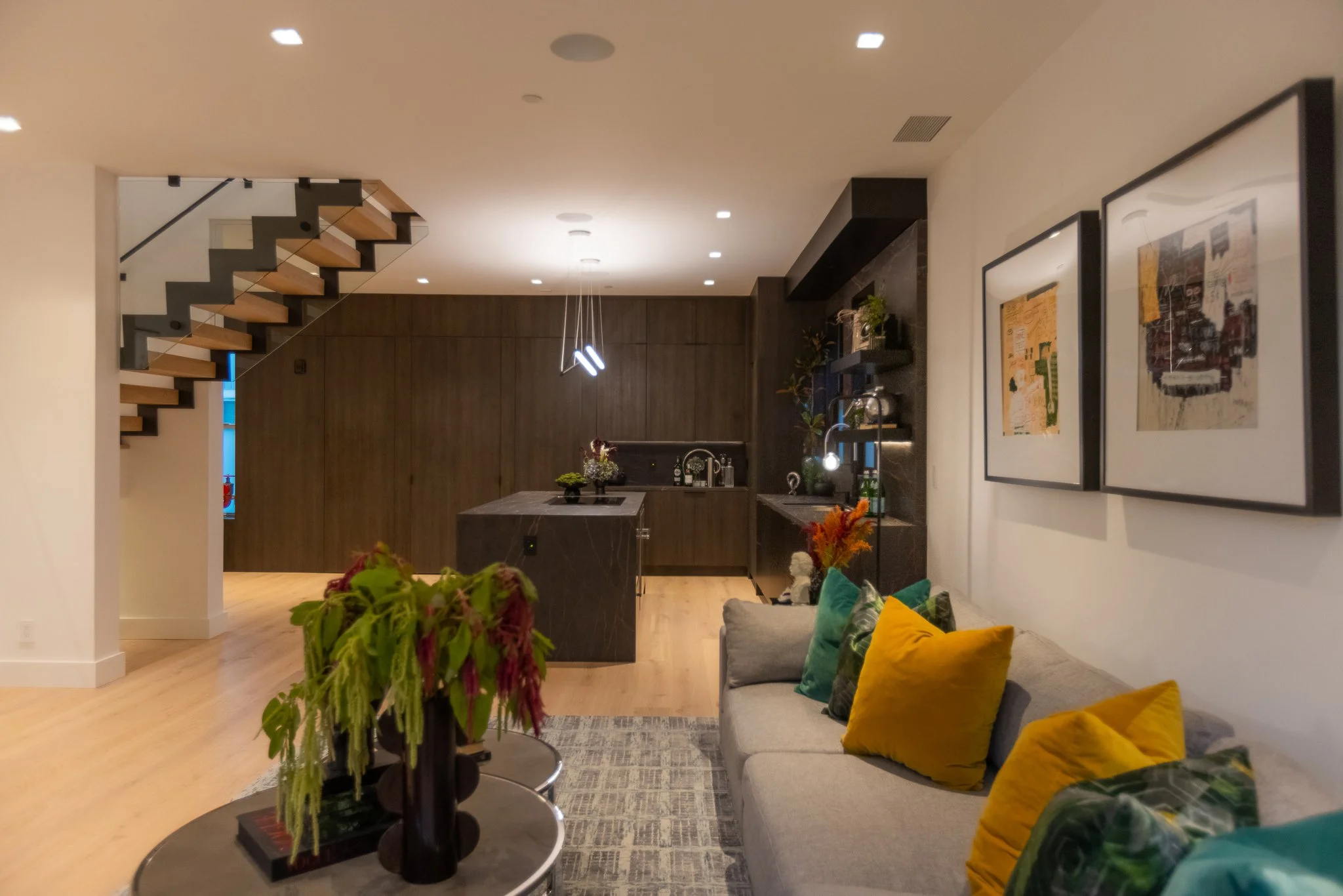 Modern living room with a staircase, a beige sofa with colorful pillows, a round coffee table with a floral arrangement, art on the wall, and a kitchen area with a dark wood island and shelves with decor.