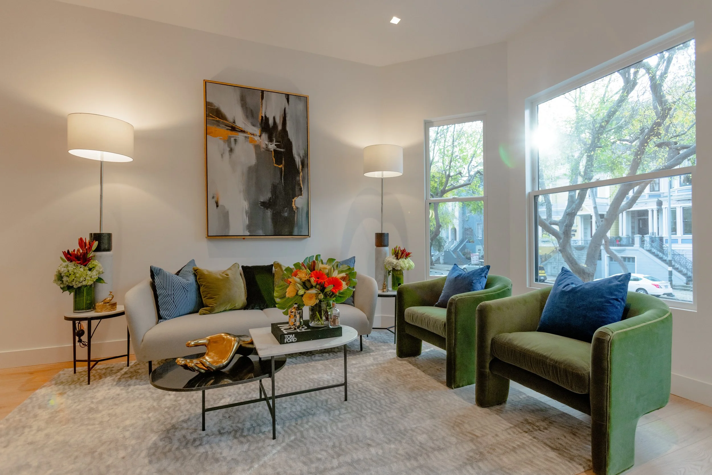 Living room with white sofa, two green armchairs, abstract painting, side tables with lamps and flowers, coffee table with a bouquet and books, large windows with trees outside.