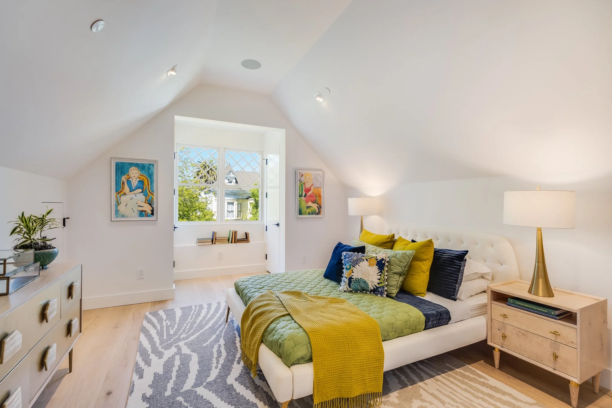 Bright bedroom with white walls and a sloped ceiling, featuring a bed with colorful pillows and a yellow blanket, wooden nightstands with lamps, a dresser with plants and decorations, and art portraits on the walls, with a window overlooking trees.