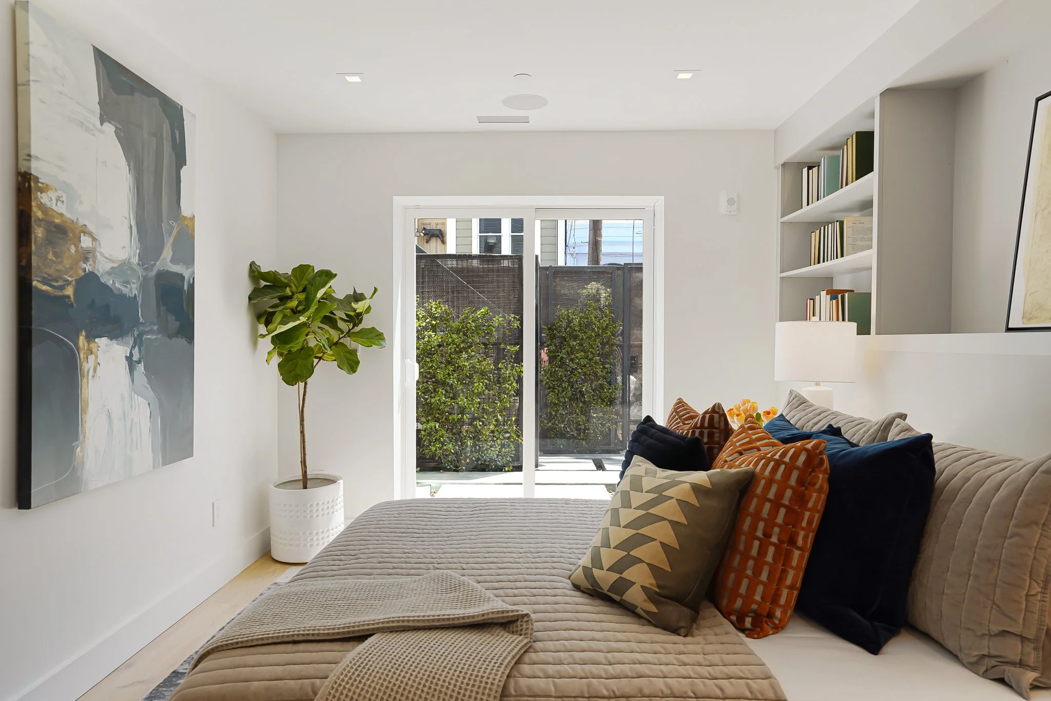 Bright bedroom with a bed, various decorative pillows, a potted plant, large artwork on the wall, a built-in bookshelf, and glass sliding door leading to an outdoor patio with greenery.