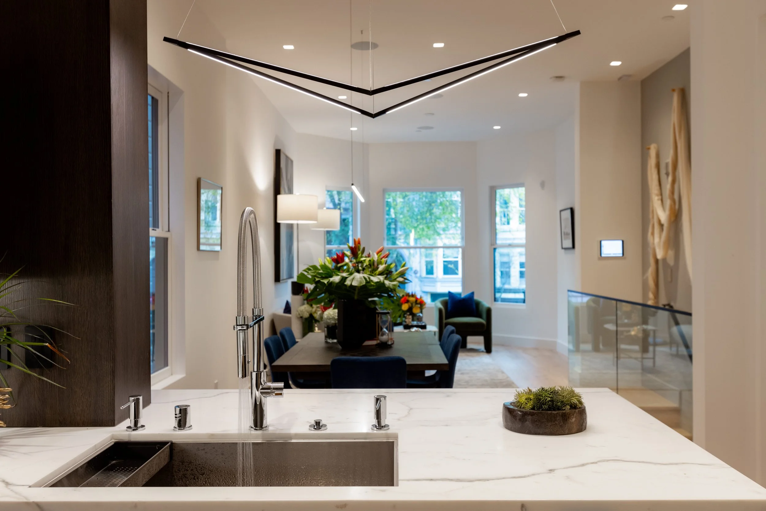 Modern kitchen with white marble countertop, black faucet, and a dining area with a vase of colorful flowers, sitting in front of large windows and a living space with armchairs and artwork.