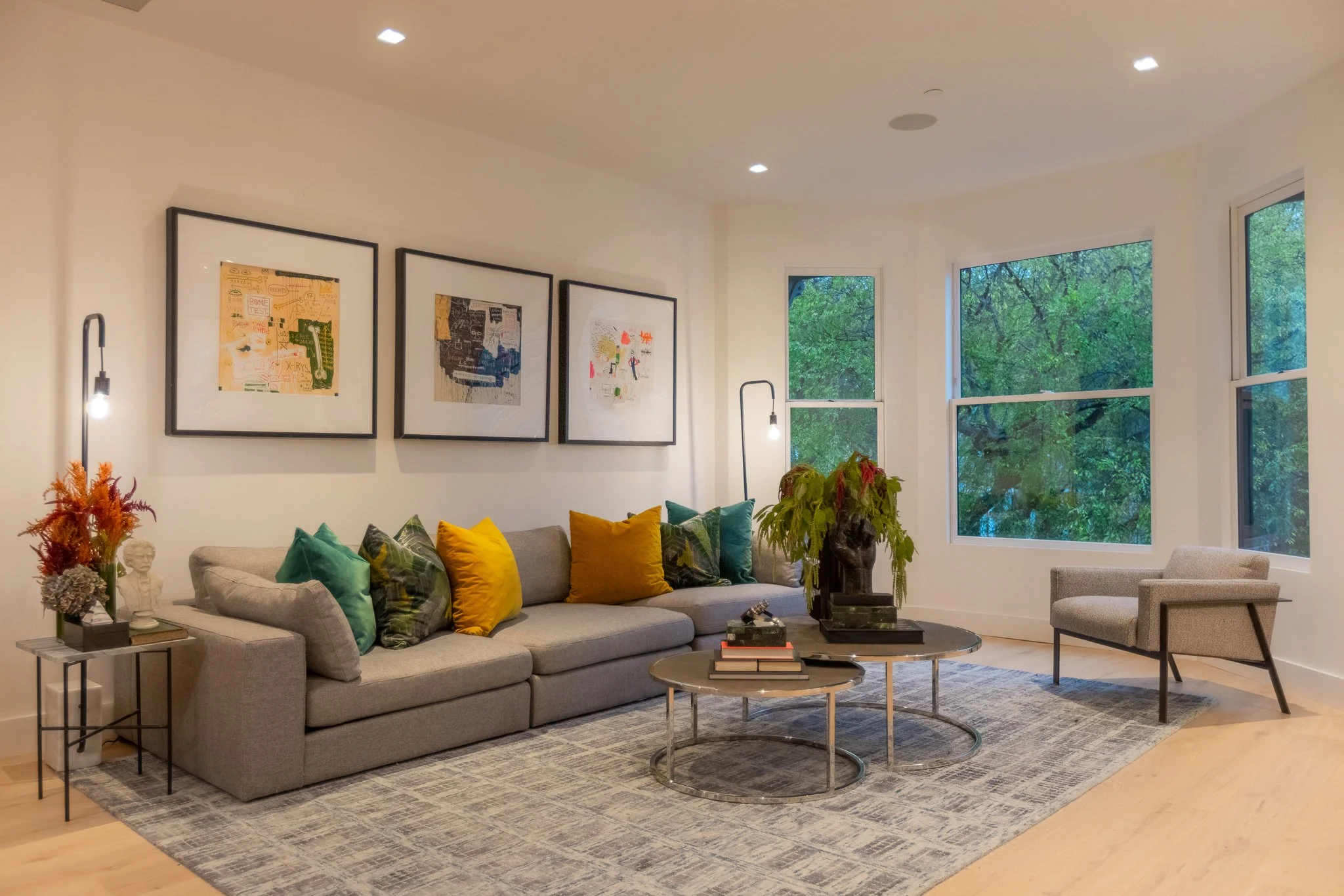 Contemporary living room with a light-colored sofa, colorful pillows, abstract artwork on the wall, a gray armchair, coffee tables, large windows showing green trees outside, indoor plants, and modern lighting fixtures.