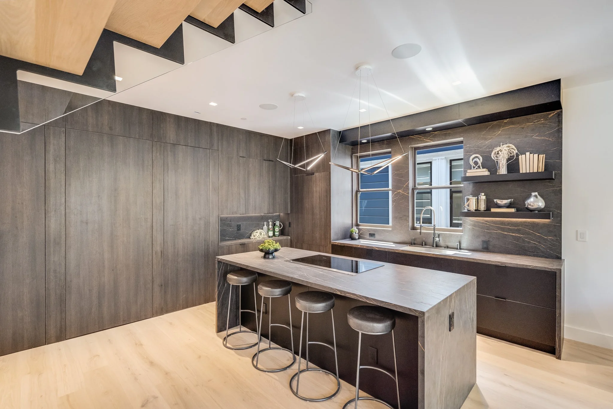 Modern kitchen with dark wood cabinetry, marble countertops, a kitchen island with four bar stools, and two windows above the sink with a view of a house exterior