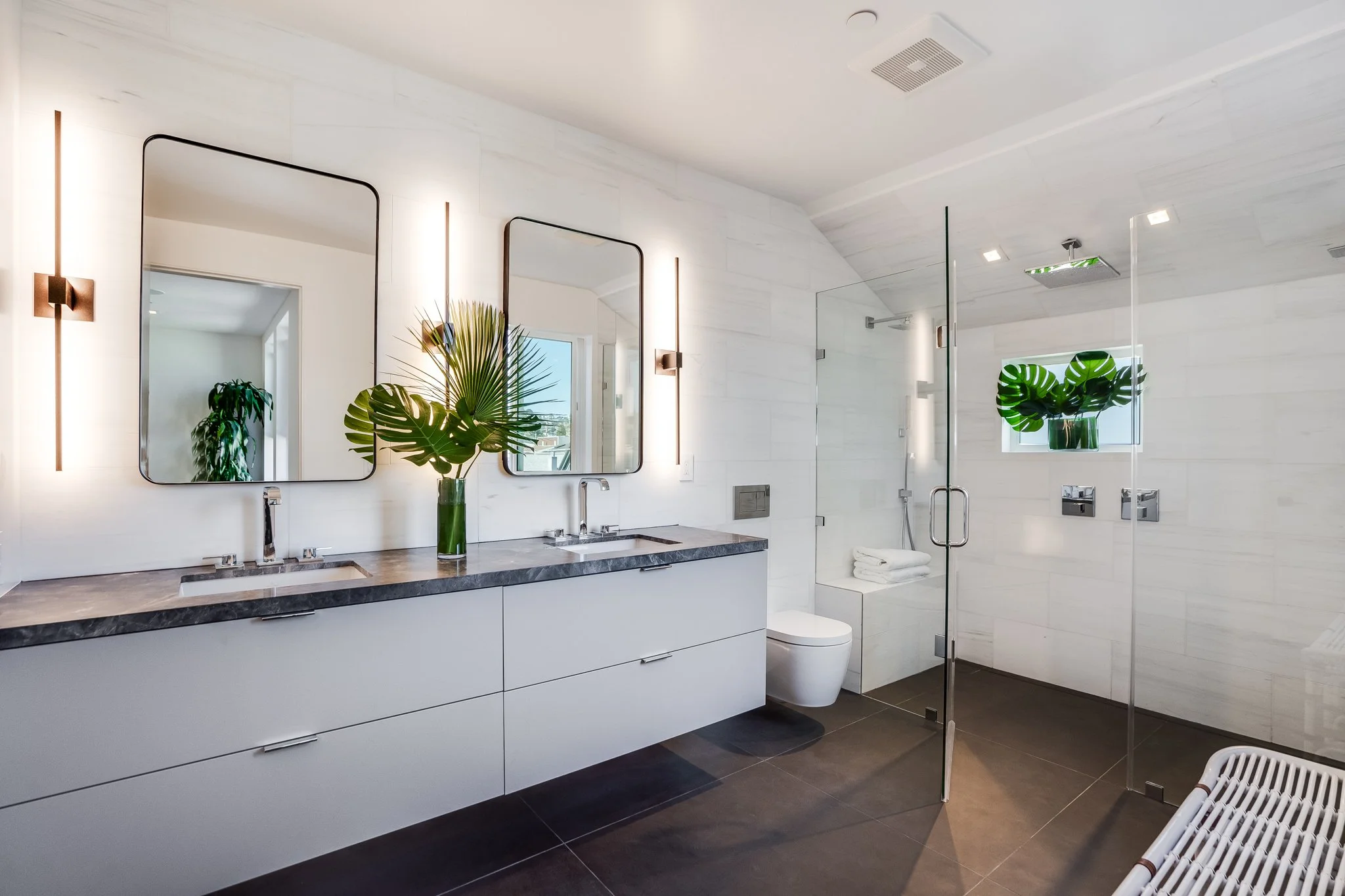 Modern bathroom with double vanity, large mirrors, pendant lights, glass shower enclosure, white walls, black floor tiles, and green plants.