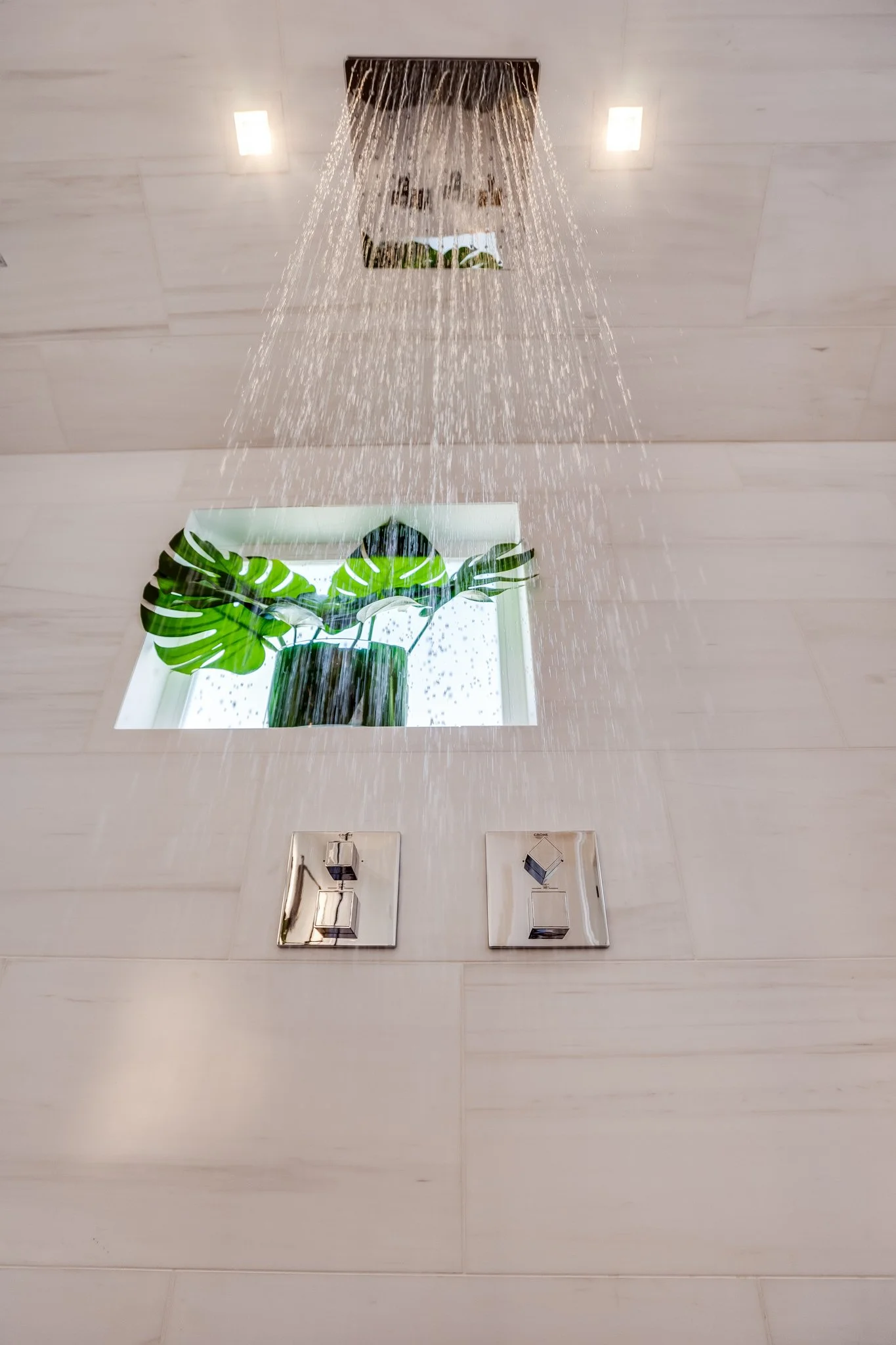 Shower with rainfall head, two switches below, white tiled wall, and a square window with a large green tropical plant visible through the window.