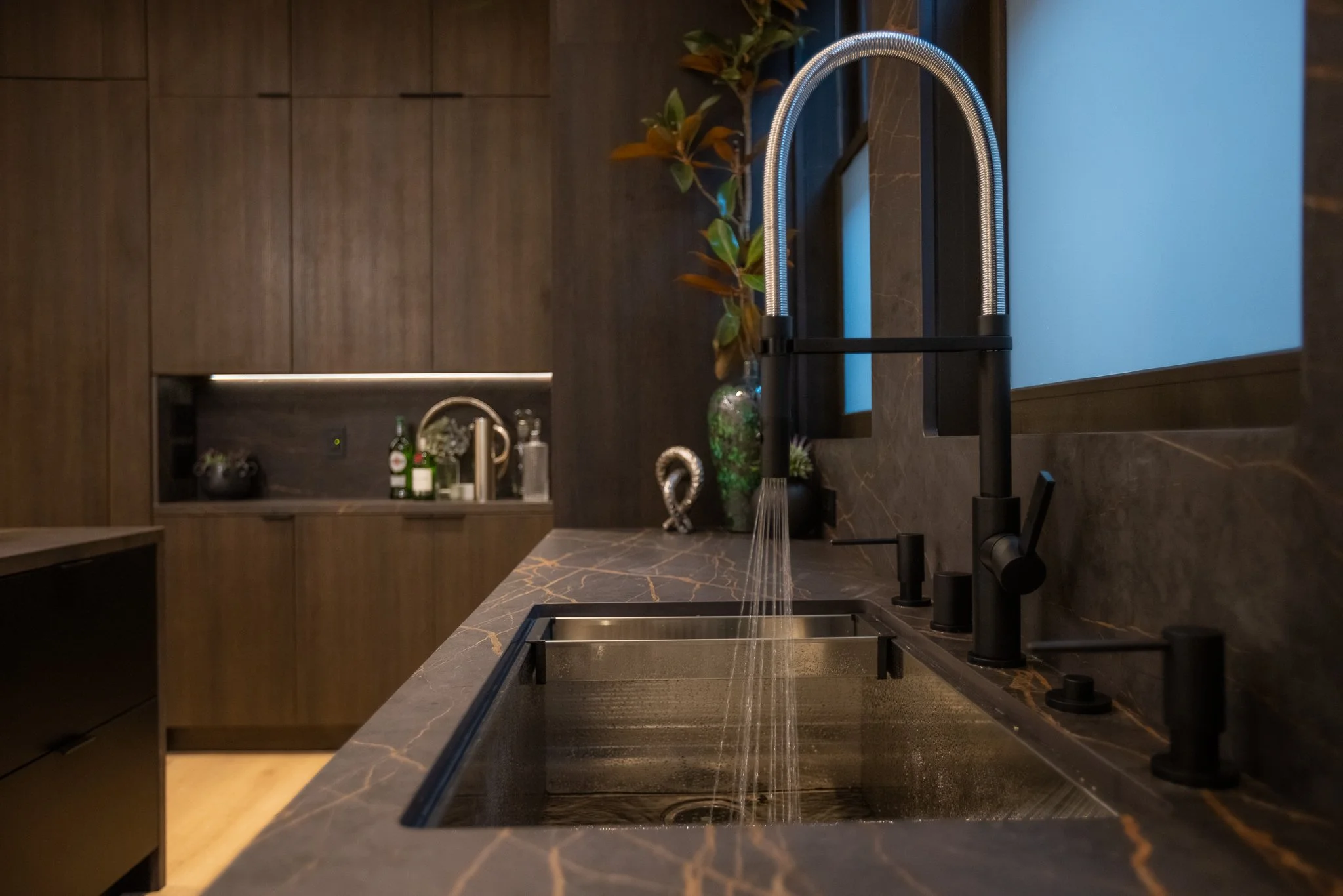 Modern kitchen with dark marble countertop and sink, black faucet, and wooden cabinets. A vase with green leaves and some decor items are on the counter, while a window is visible in the background.