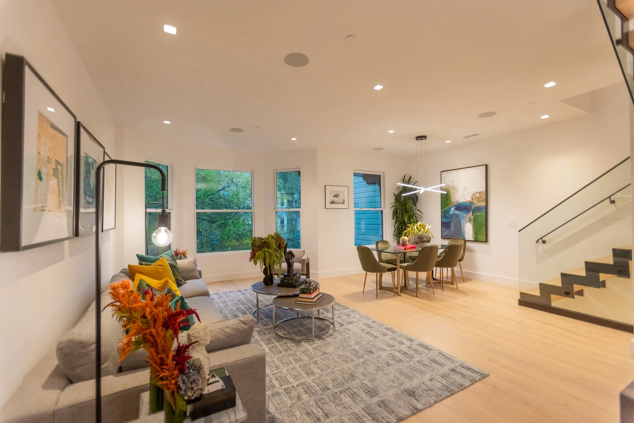 Open living and dining room with large windows, chandelier, artwork, gray sofa with colorful pillows, round coffee tables, plants, and a staircase with black steps.