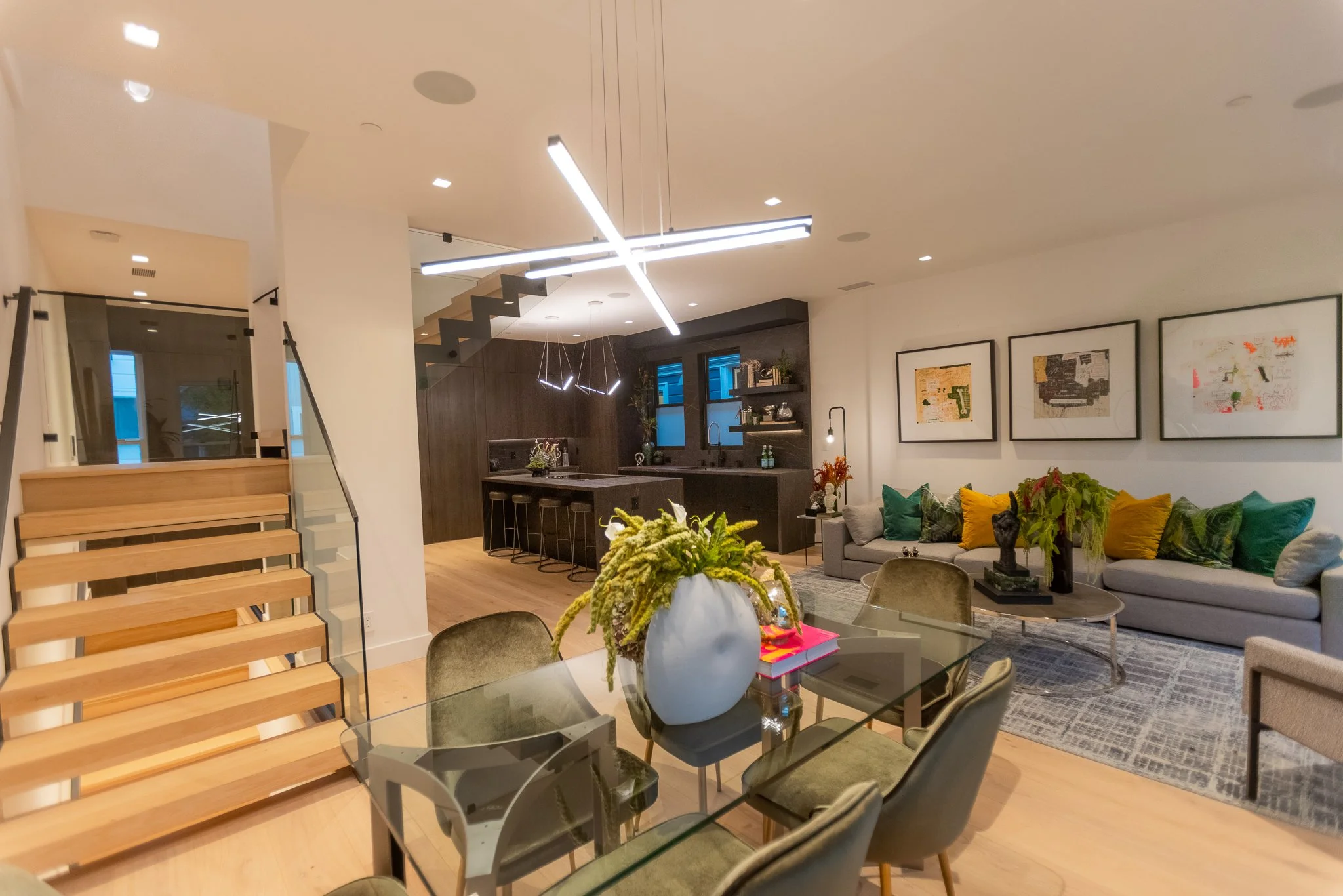 Open-concept living room and kitchen area with modern decor, a sectional sofa with colorful cushions, framed artwork on the wall, a glass dining table with a large vase of flowers, and a wooden staircase leading upstairs.