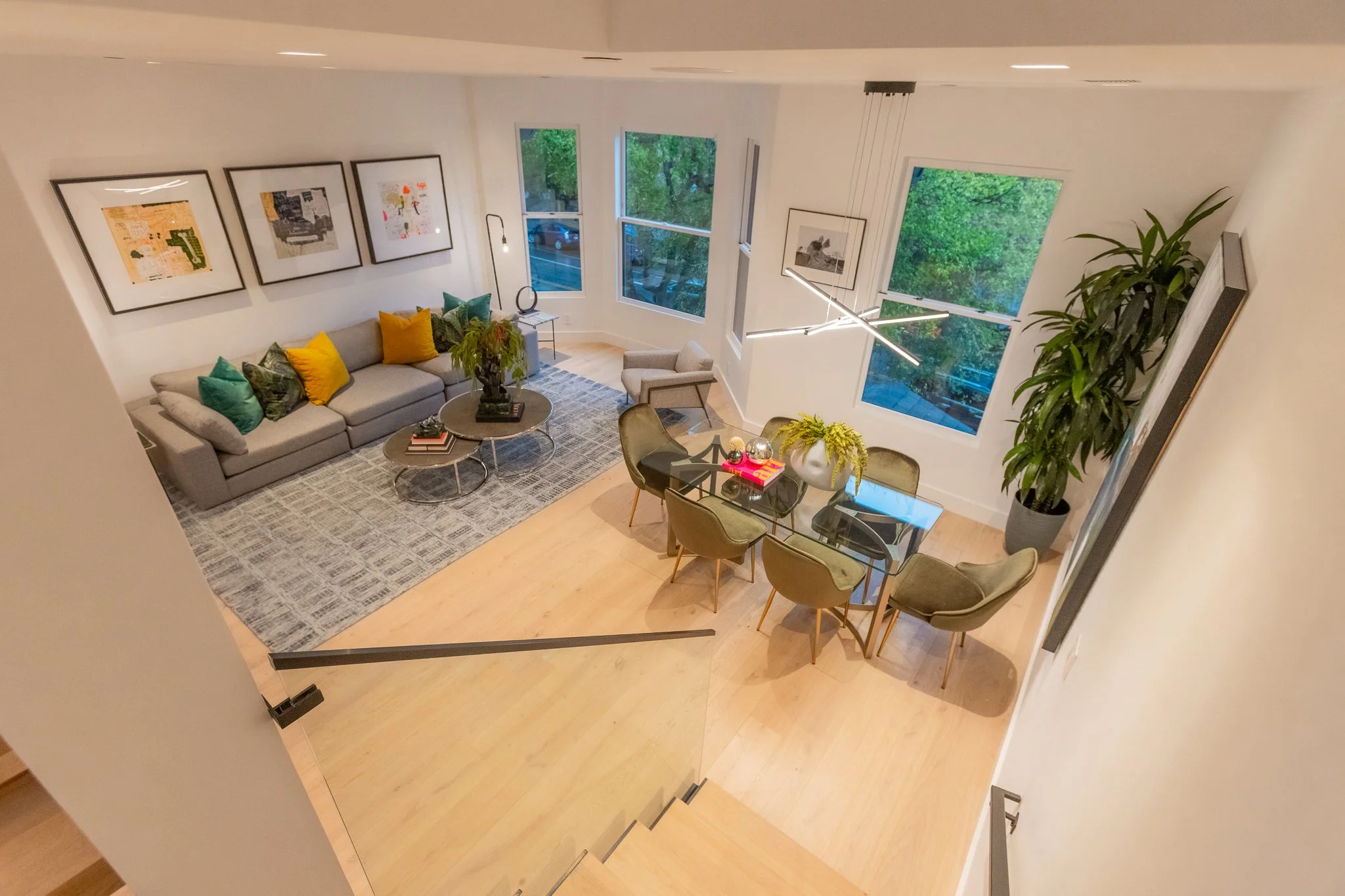 A modern living and dining area with large windows, a light gray sofa with colorful pillows, a gray armchair, a round coffee table, a glass dining table with green chairs, interior decorations including framed artwork, plants, and a contemporary chan