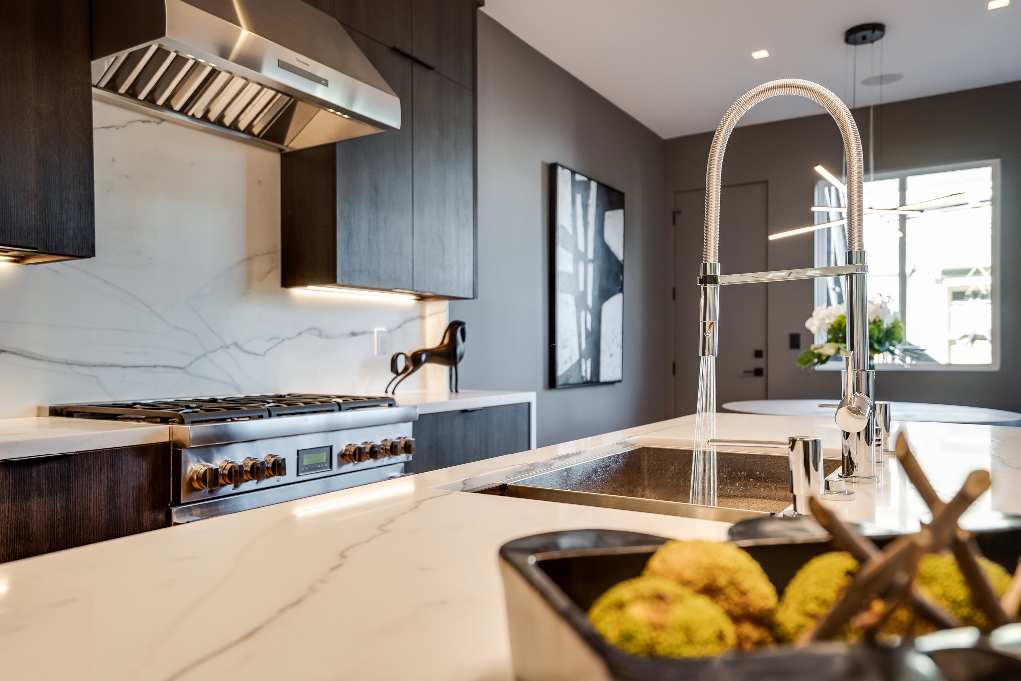 Modern kitchen with marble countertops, dark wood cabinets, stainless steel range hood, and a sink with a flexible faucet. There is a decorative horse figure on the counter and a basket of green apples in the foreground, with a window and artwork on 