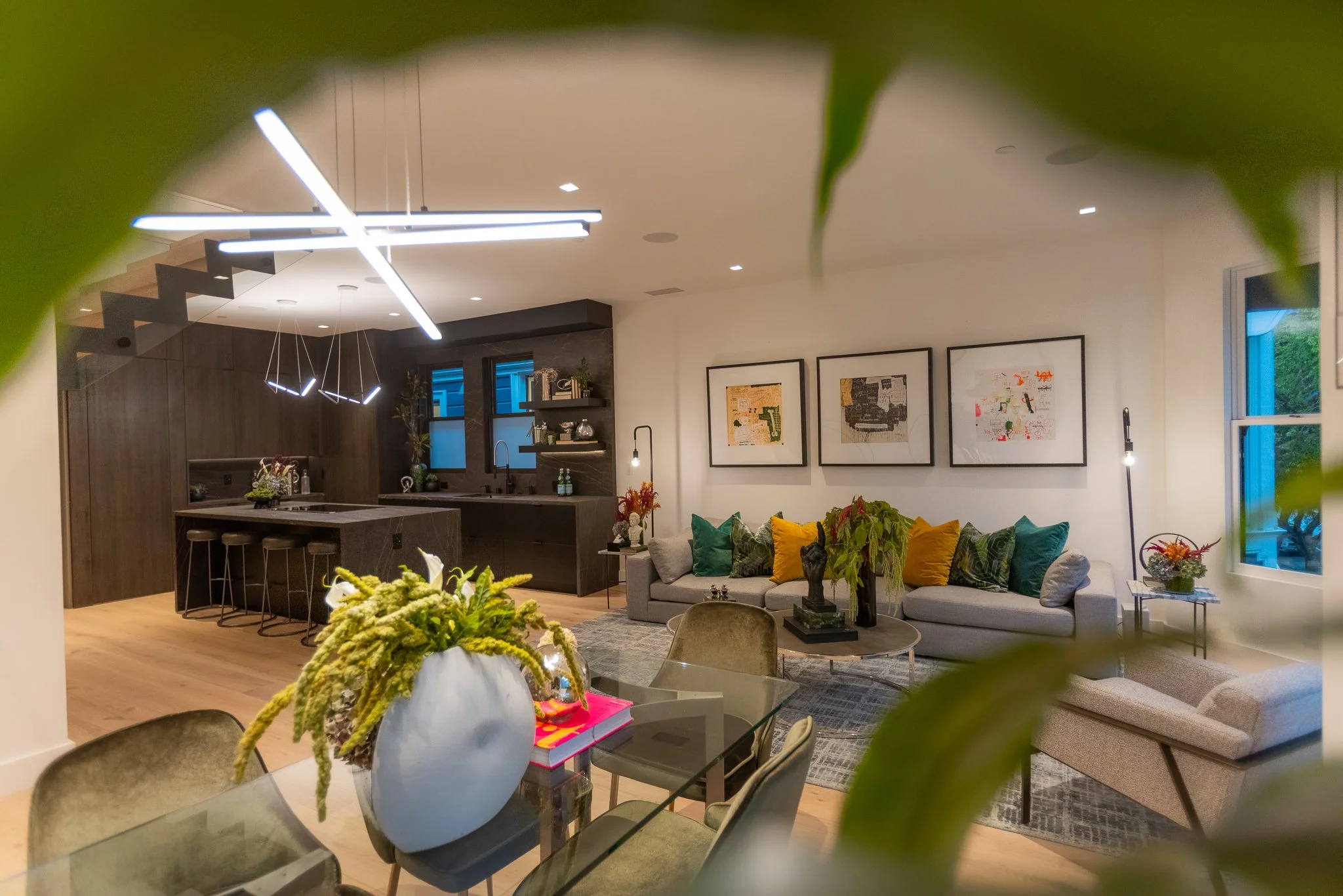 Modern living room and kitchen with colorful pillows, abstract art, and contemporary lighting fixtures, seen through leafy foreground.