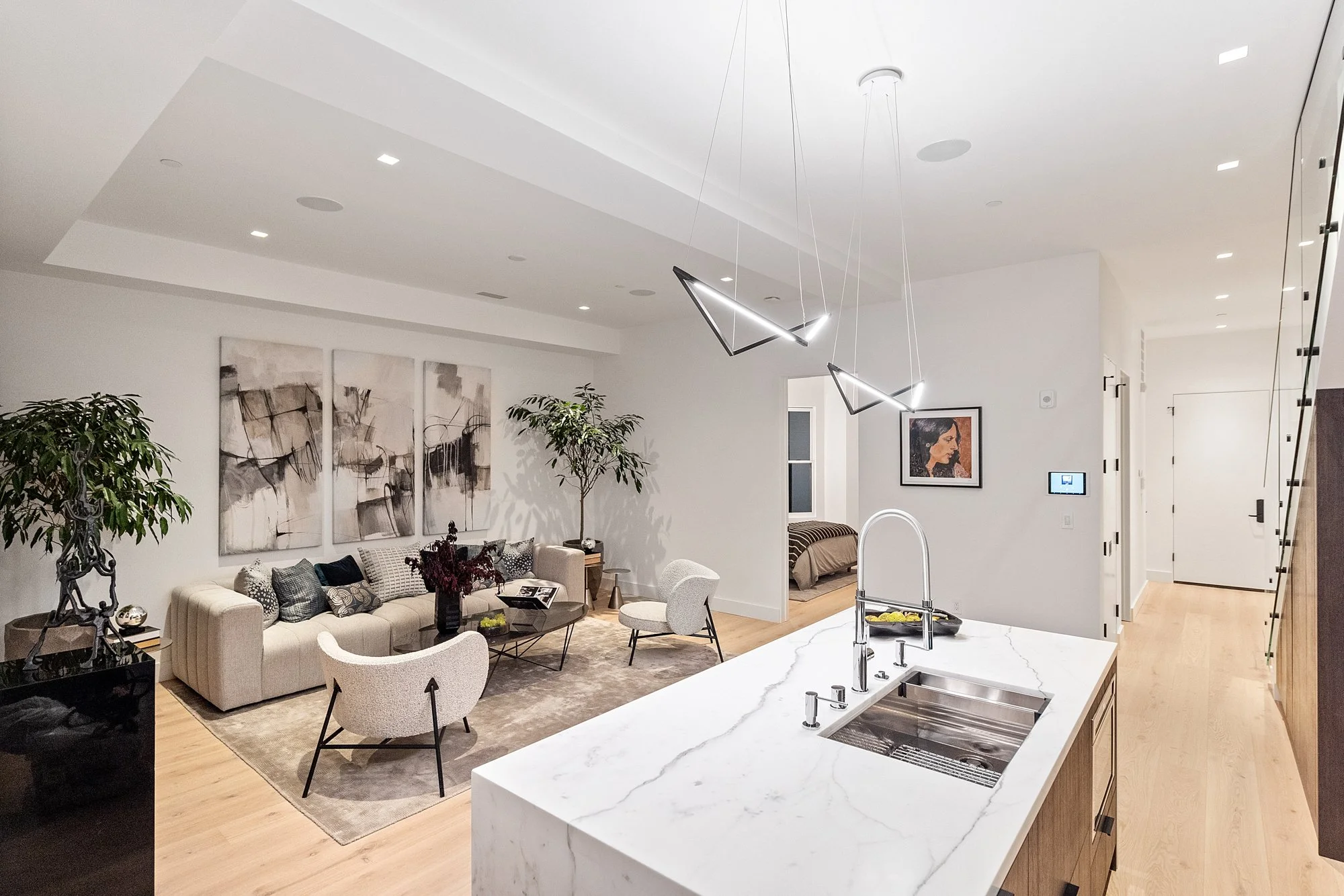 Modern open-concept living room and kitchen with white walls, wooden floors, and contemporary furnishings, including a beige sofa, armchairs, large abstract artwork, and pendant lighting fixtures.