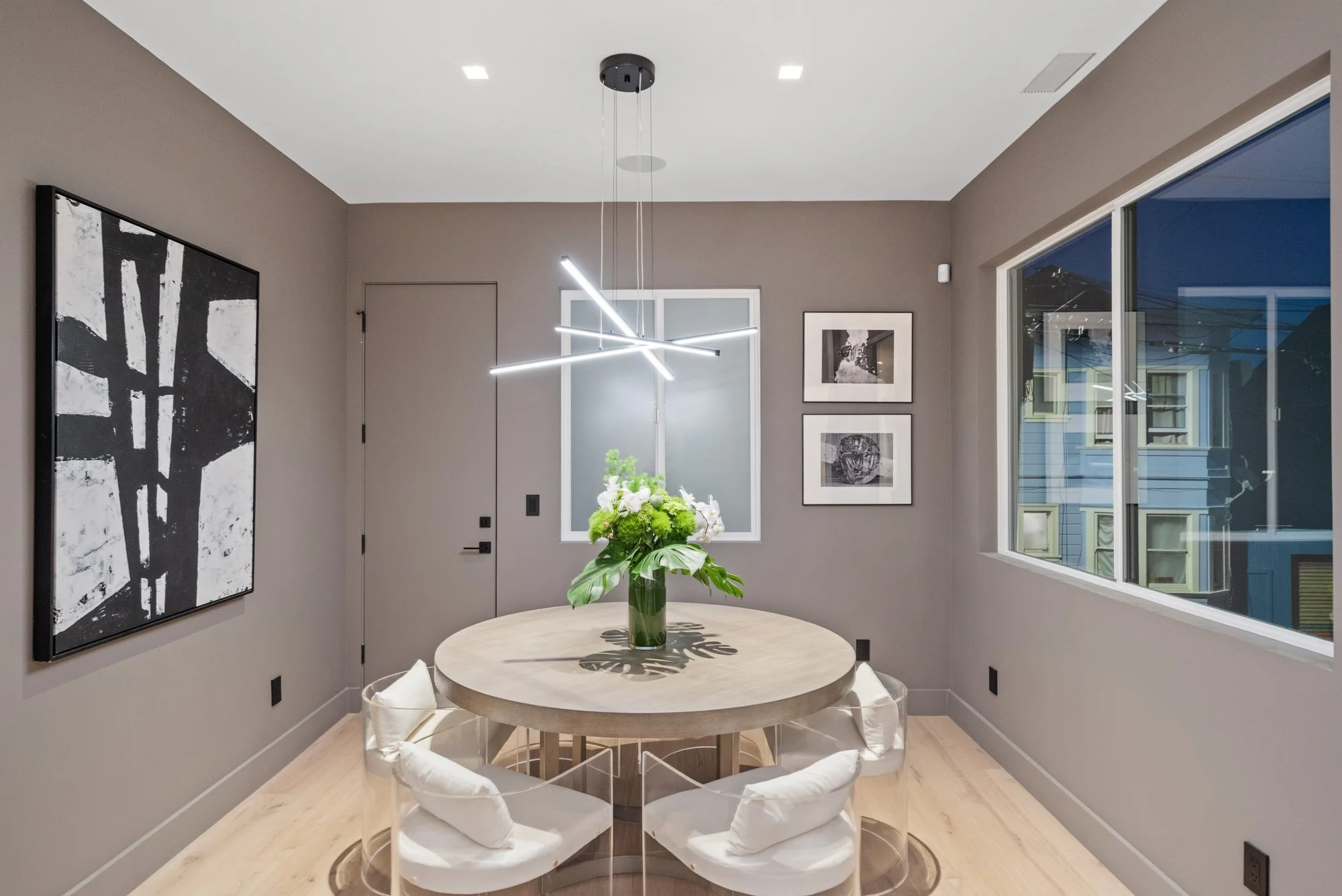 Modern dining room with a round wooden table, glass chairs with white cushions, and a geometric LED chandelier, with wall art and large windows showing a nighttime exterior.