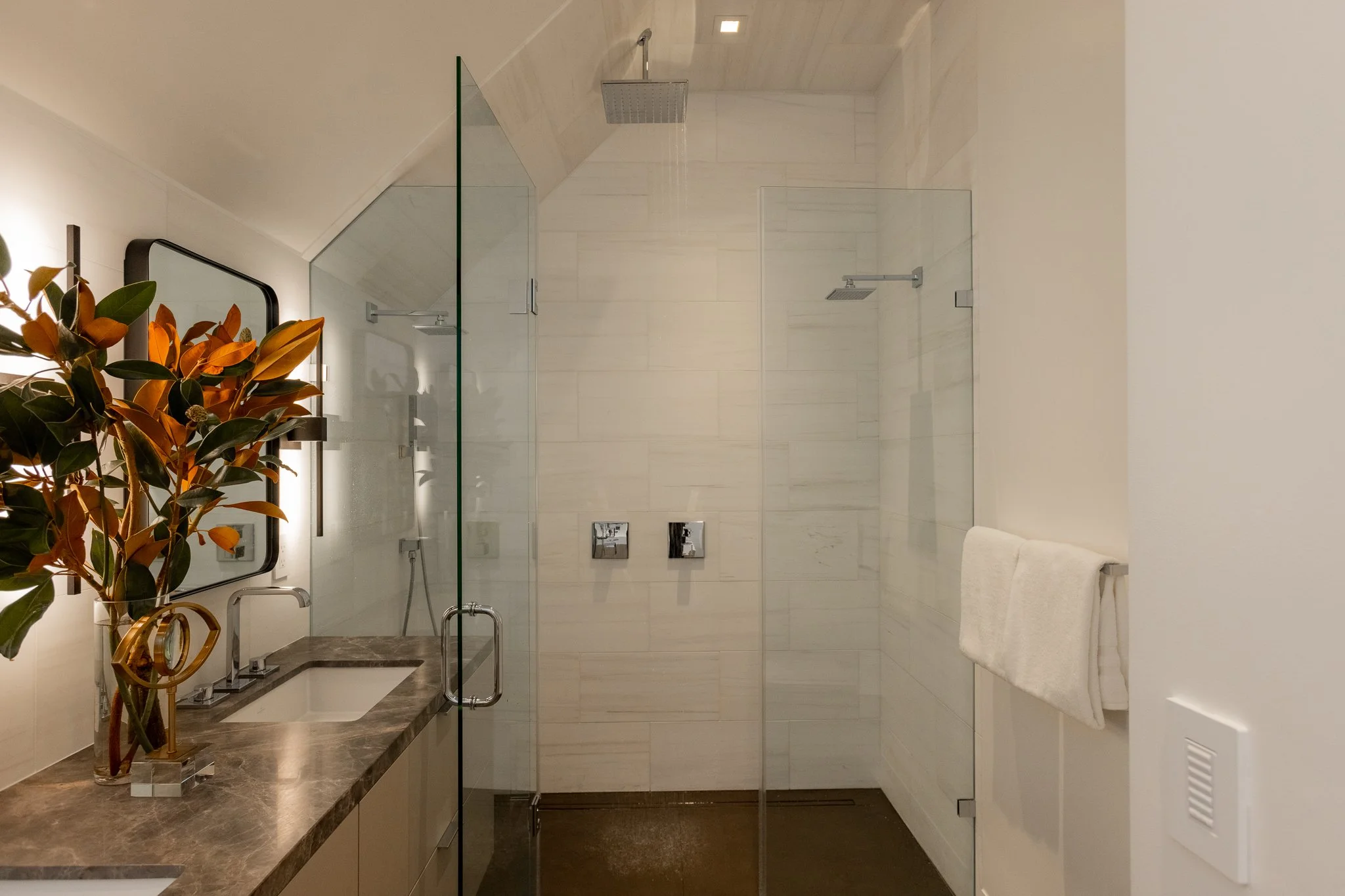 Modern bathroom with a glass-enclosed shower, white tiling, a marble countertop with a sink, a large mirror, and a towel hanging on a rack.