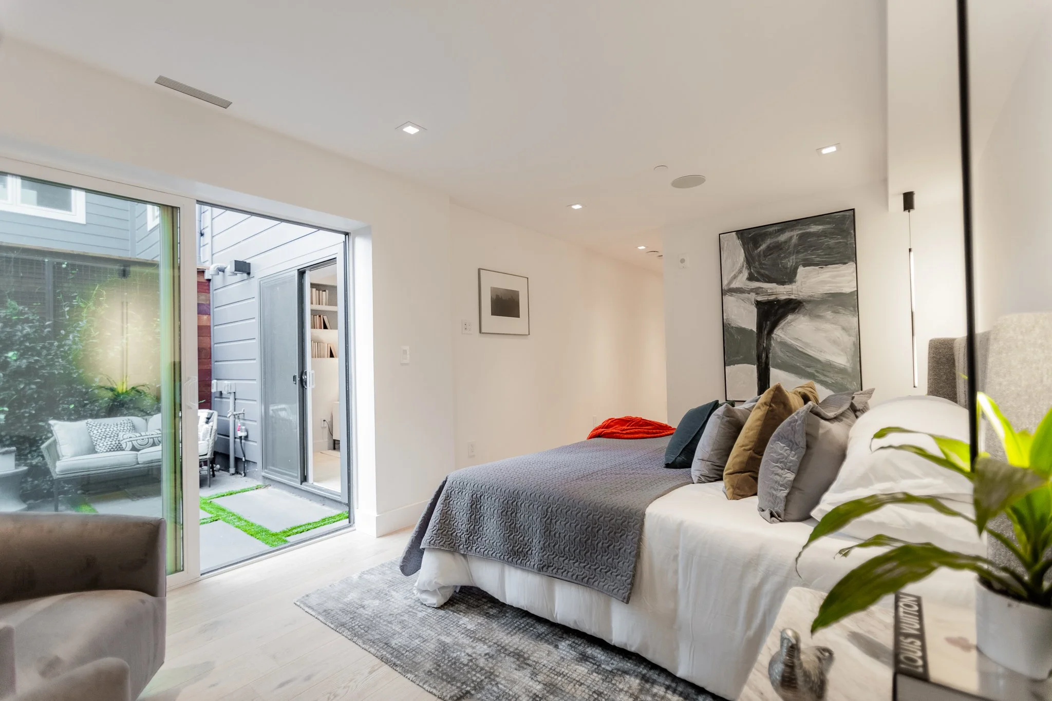 Modern bedroom with a bed covered in gray and white bedding, colorful pillows, and a large black and white abstract painting on the wall. A sliding glass door opens to a small outdoor patio with outdoor seating and greenery.