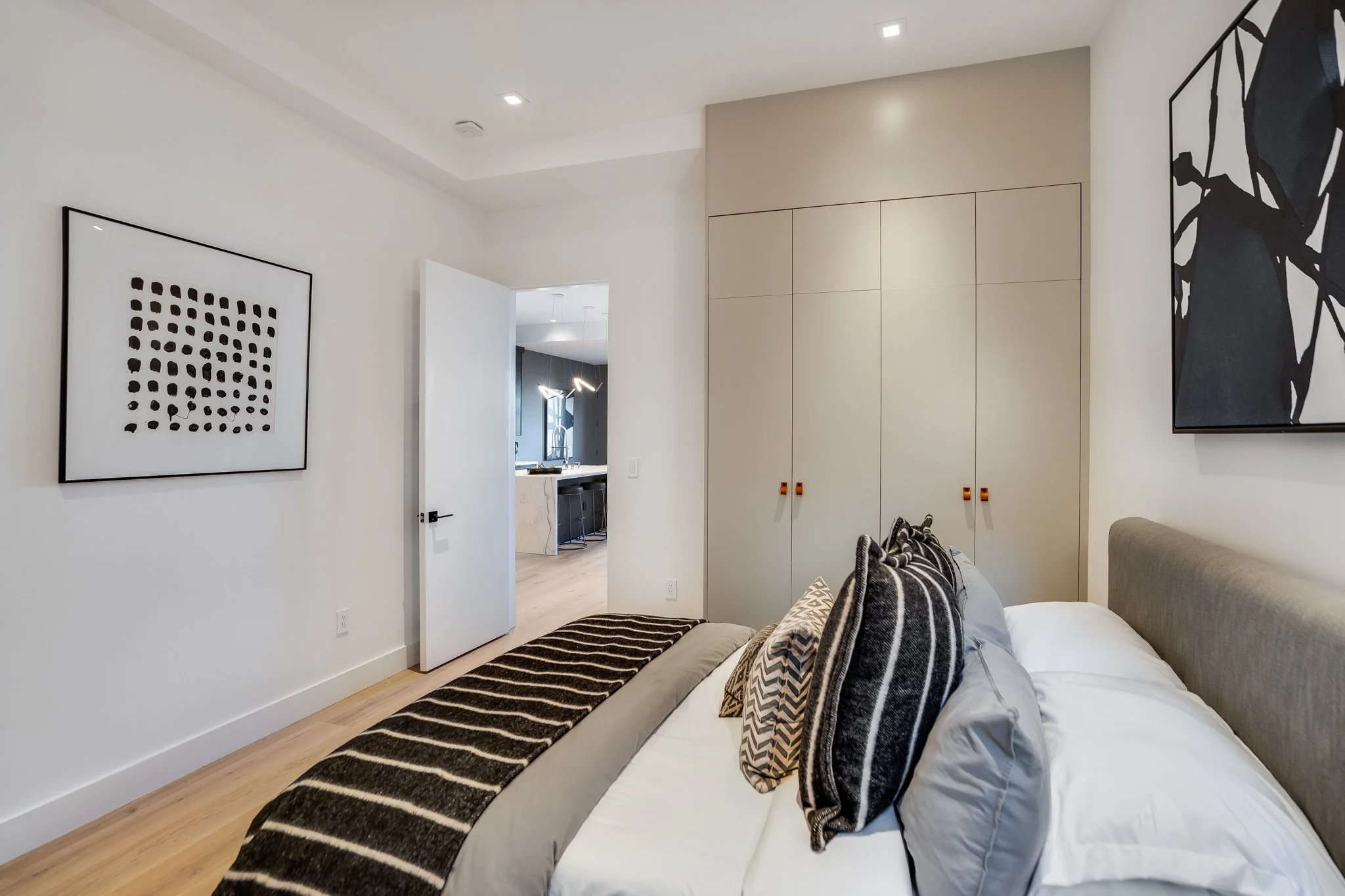 A bedroom with white walls and a bed with multiple pillows, a beige headboard, and a black and white striped blanket, adjacent to a large beige closet, art on the walls, and an open door leading to a kitchen area.