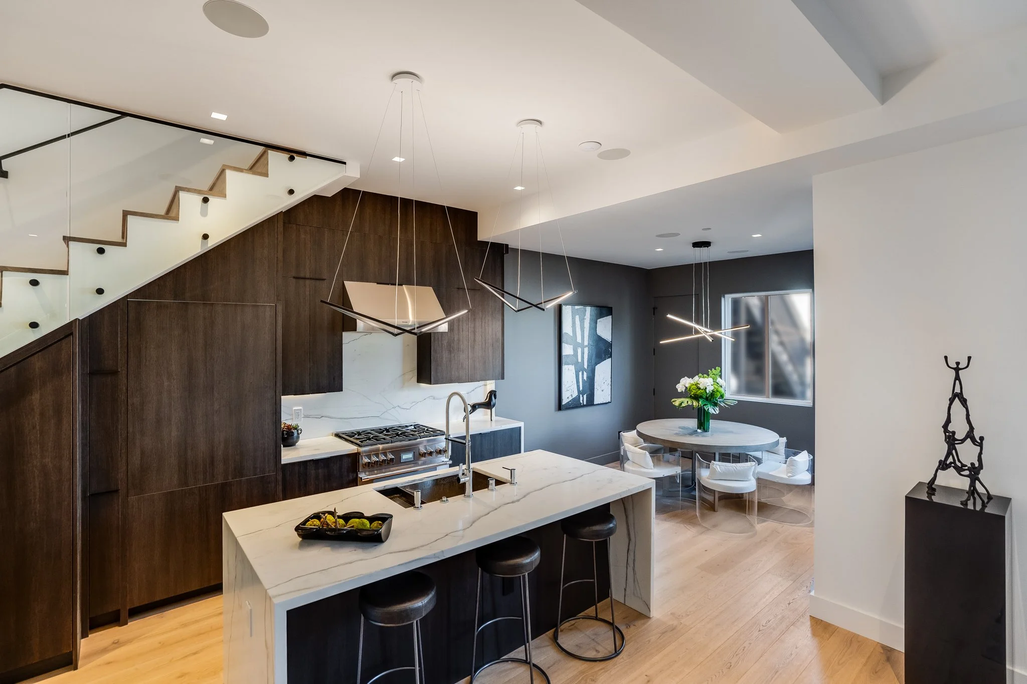 Modern open-concept kitchen with dark wood cabinetry, white marble island, stainless steel appliances, and a round dining table with white chairs and a flower centerpiece.