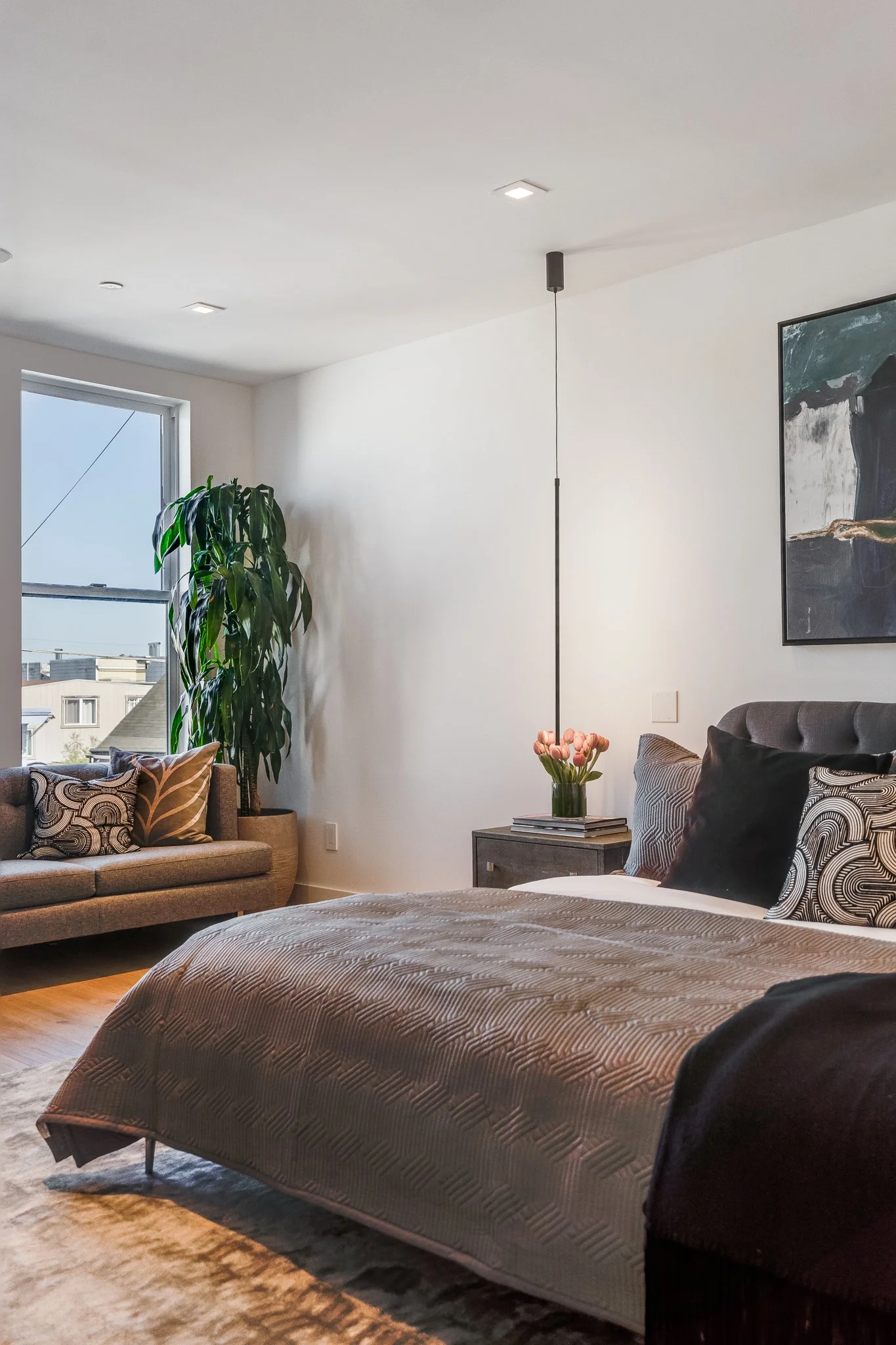 A bedroom with a gray bed, black and patterned pillows, a small side table with a vase of pink tulips, a large plant near a window, a sofa with matching pillows, and modern artwork on the wall.