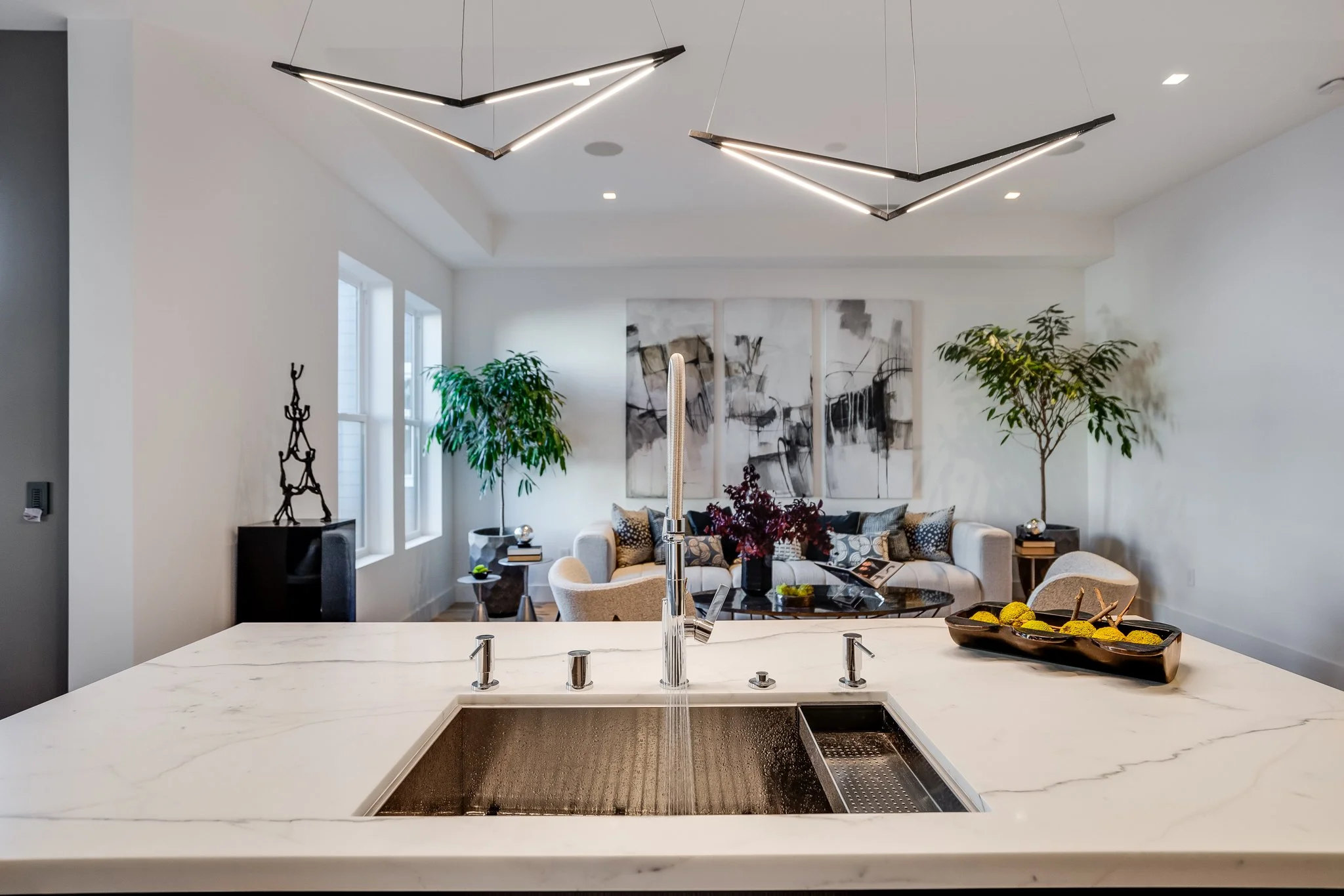 Modern kitchen island with a marble countertop and sink, overlooking a living room with modern decor, including a white sofa, abstract art, and green potted plants.