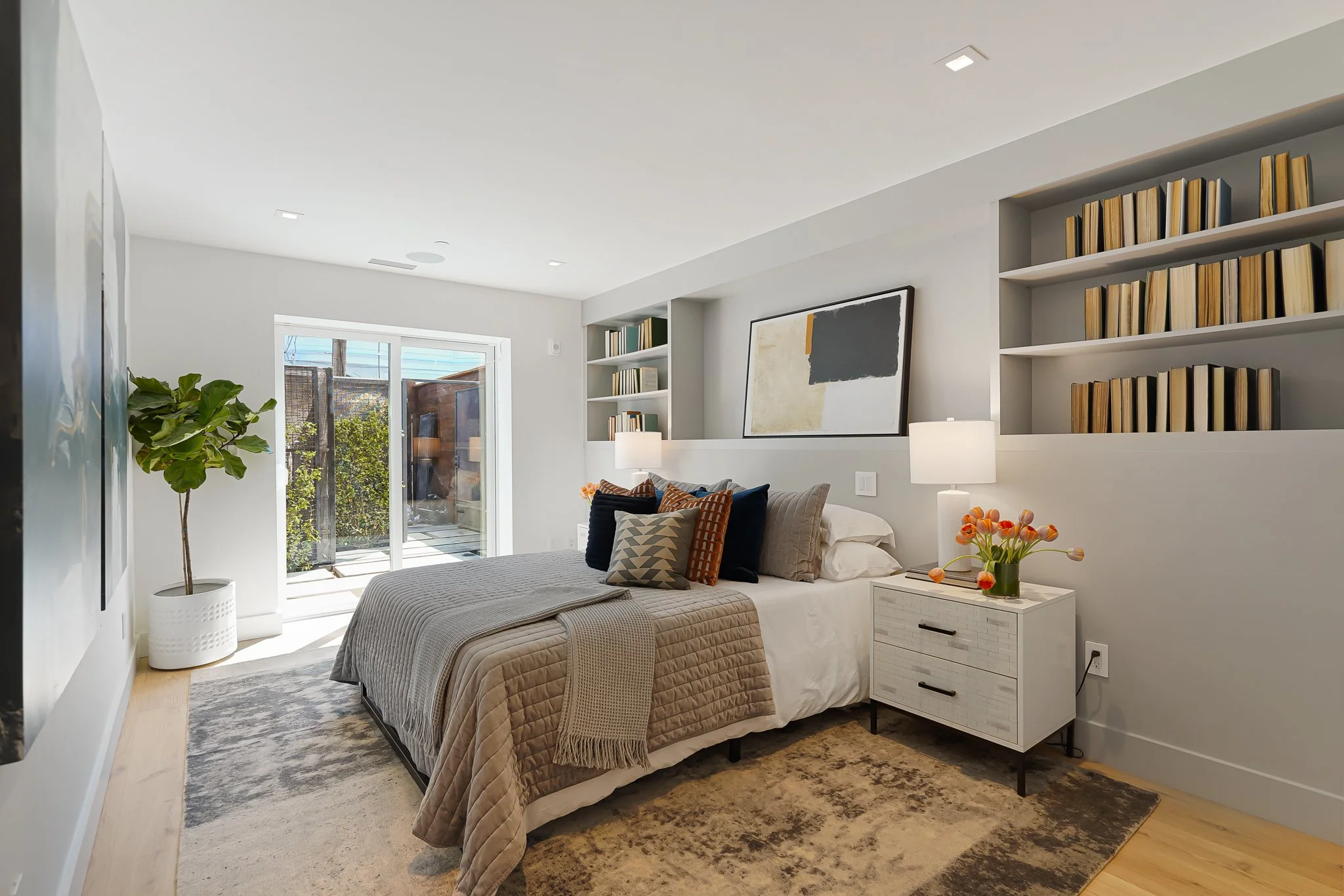 Modern bedroom with bed, pillows, nightstand with flowers, bookshelf, artwork, sliding glass door, and potted plant.