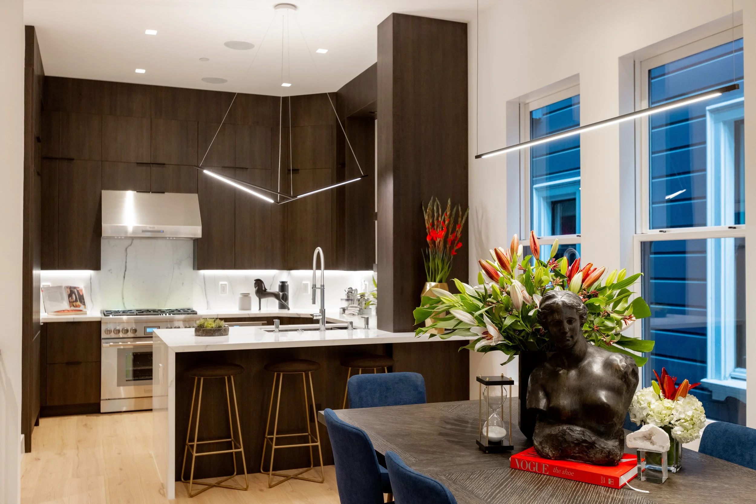 Modern kitchen with dark wood cabinets, white marble backsplash, and a white island with bar stools. A dining table in the foreground has a large floral arrangement, a bust sculpture, a clock, and a red book. Large windows show blue evening exterior.