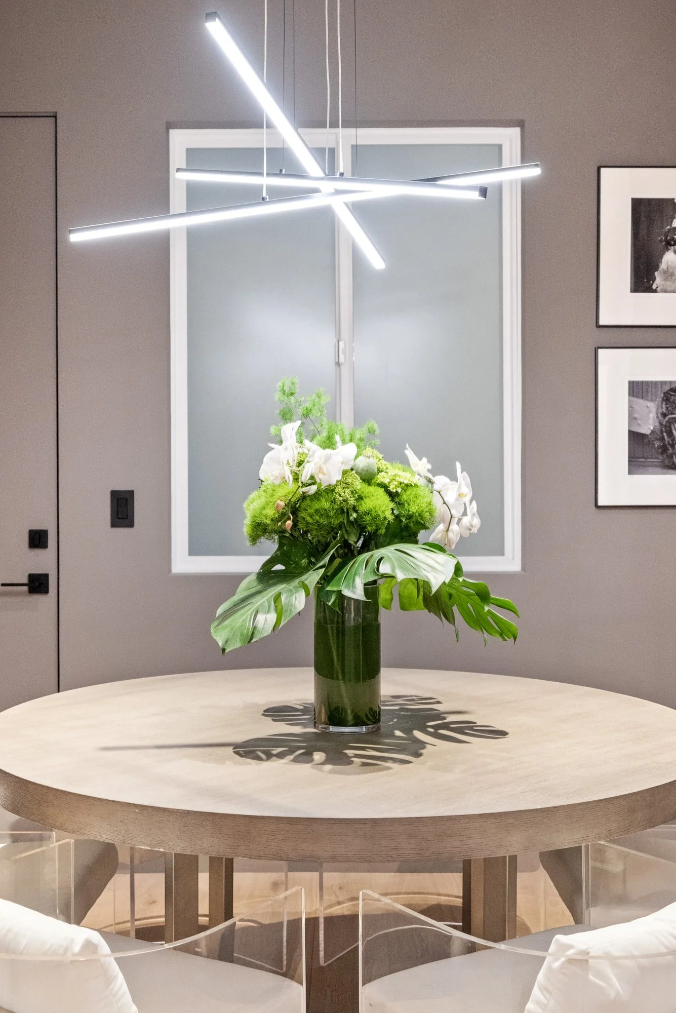 A flower arrangement in a tall glass vase on a round wooden table with modern overhead lighting above.